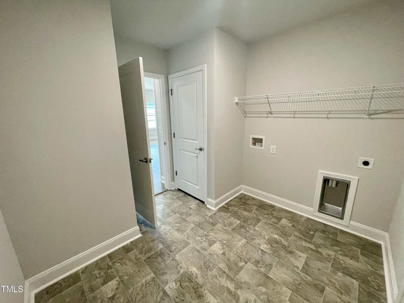 A laundry room with light gray walls, tiled floor, two closed white doors, a wire shelf on the right wall, and hookups for a washer and dryer below the shelf.