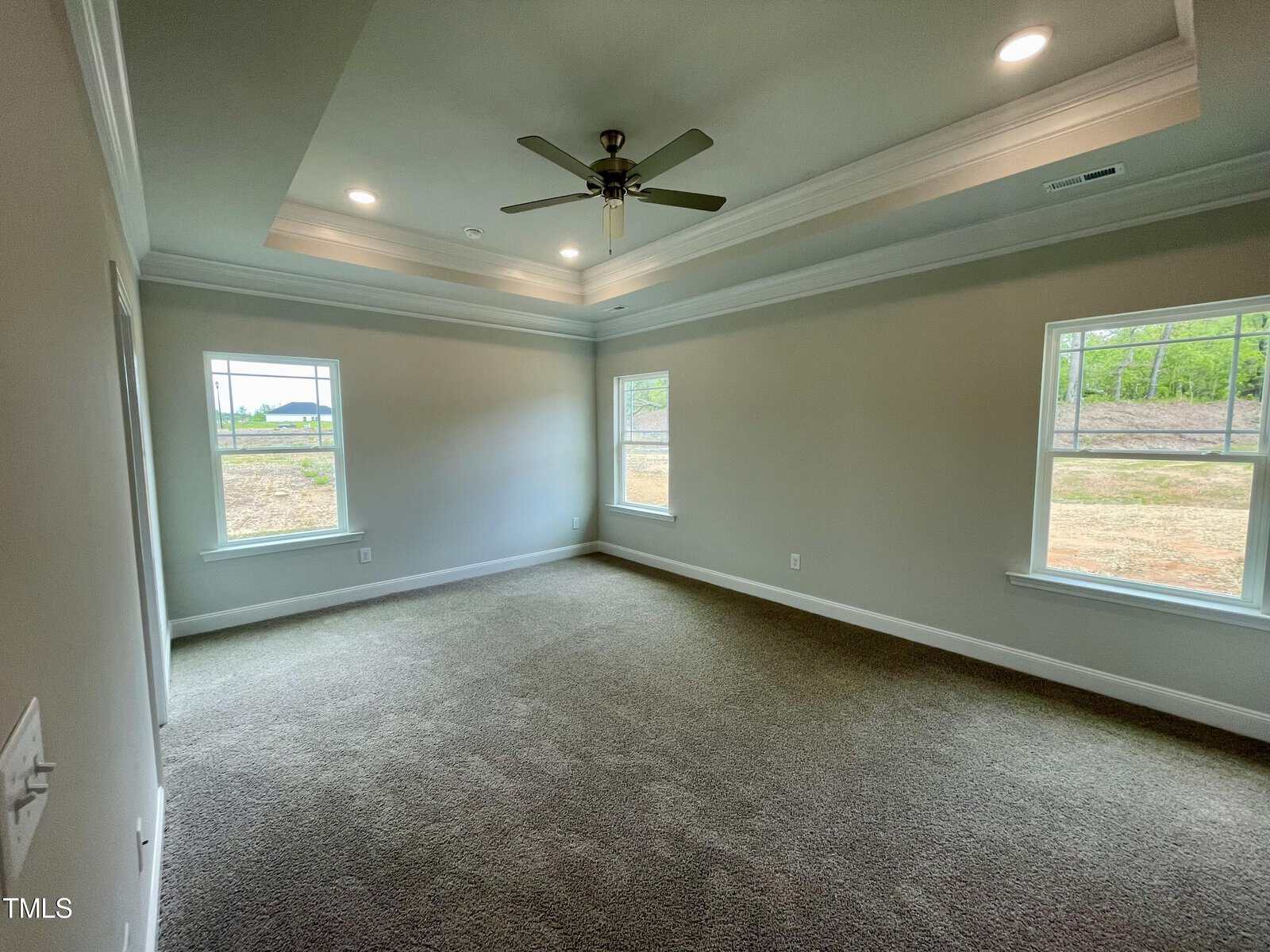Spacious empty room with beige carpet, light gray walls, tray ceiling with recessed lights, ceiling fan, and three large windows letting in natural light.