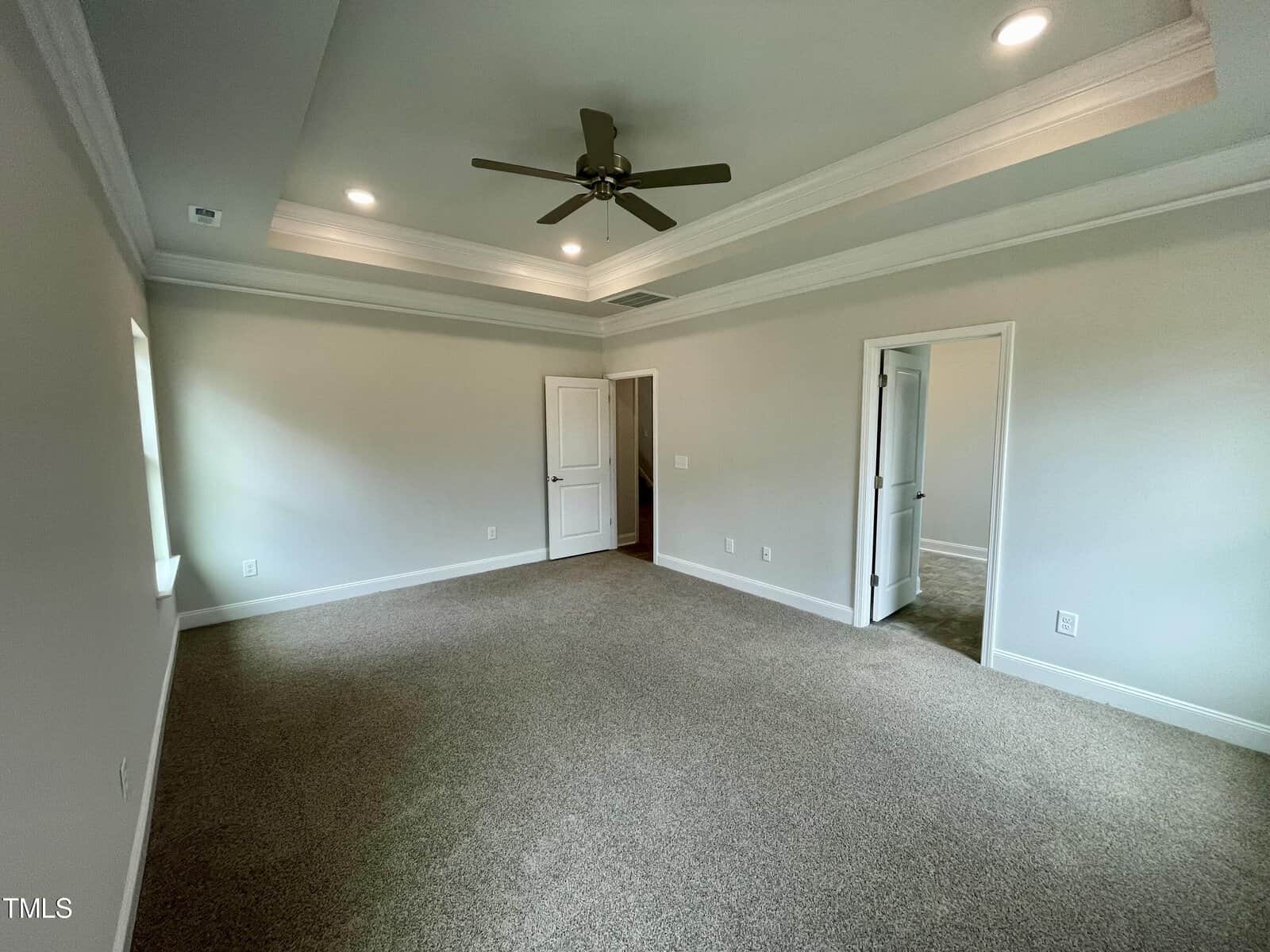 Empty bedroom with beige walls, carpeted floor, tray ceiling with recessed lighting, ceiling fan, and two doors—one closed, one open—leading to another room with similar carpet. Natural light enters from a window on the left.