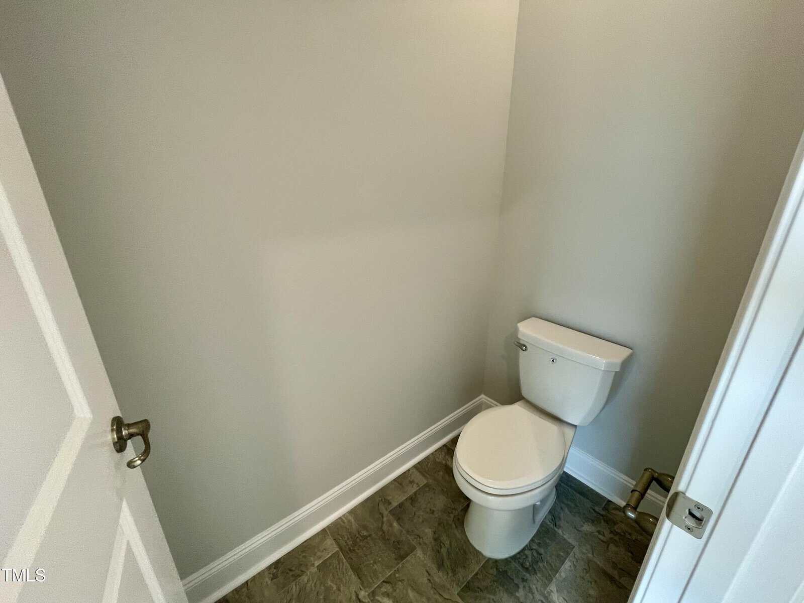 A small bathroom with a white toilet, gray tile floor, light beige walls, and a door partially open on the left.