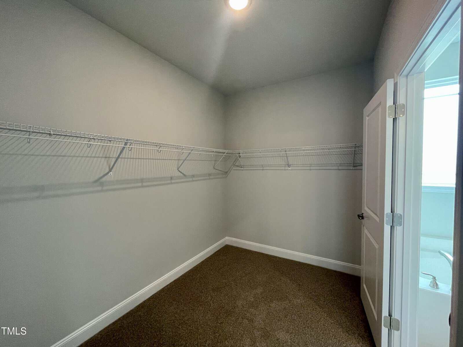 Empty walk-in closet with beige carpet, gray walls, white baseboards, and wire shelving along two walls. An open door on the right leads to a brightly lit bathroom. Ceiling light fixture is on.