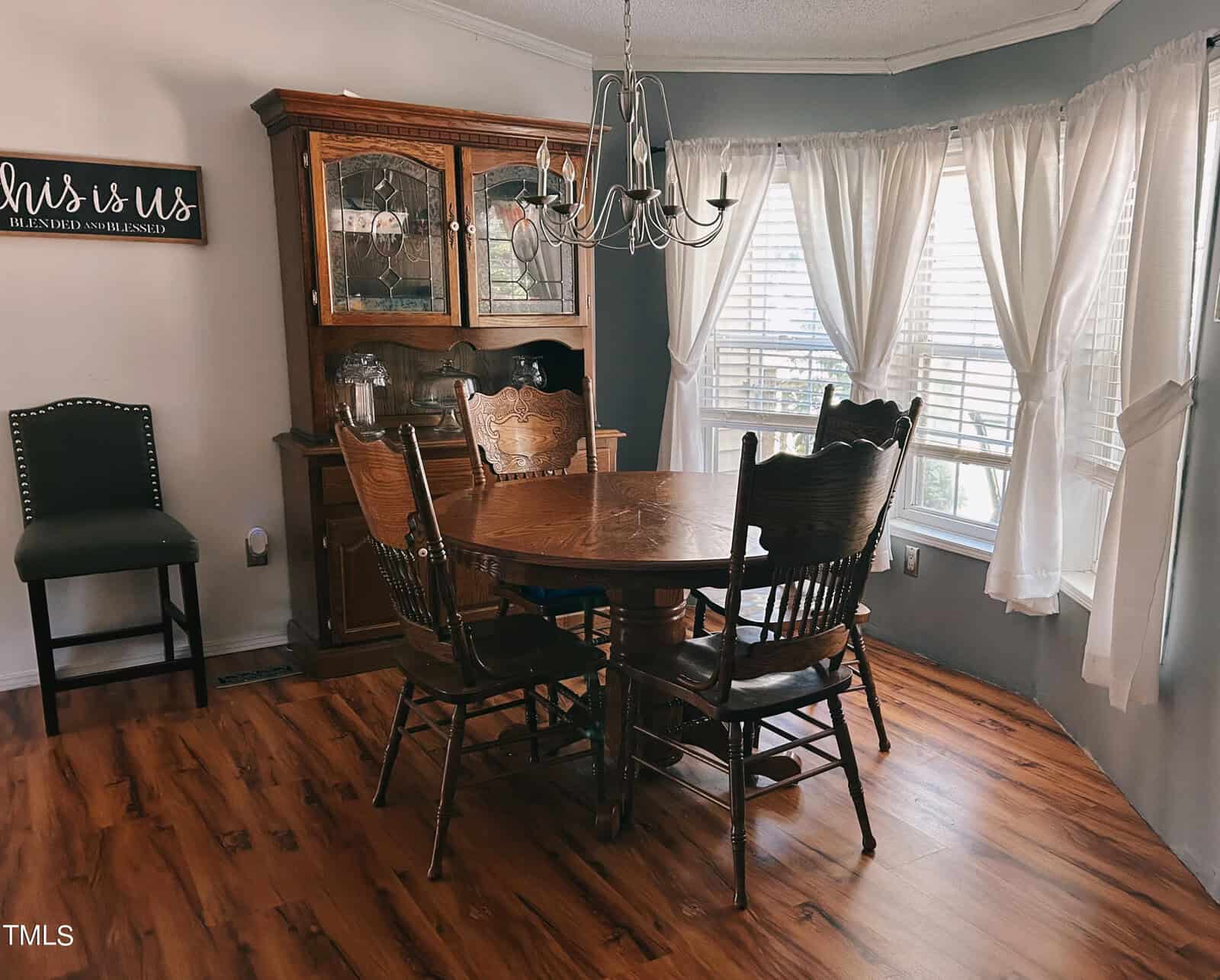 A dining room with a round wooden table and five chairs on a hardwood floor. A wooden hutch stands in the corner. A large window with sheer curtains lets in natural light. A wall sign reads "this is us" above an upholstered chair.