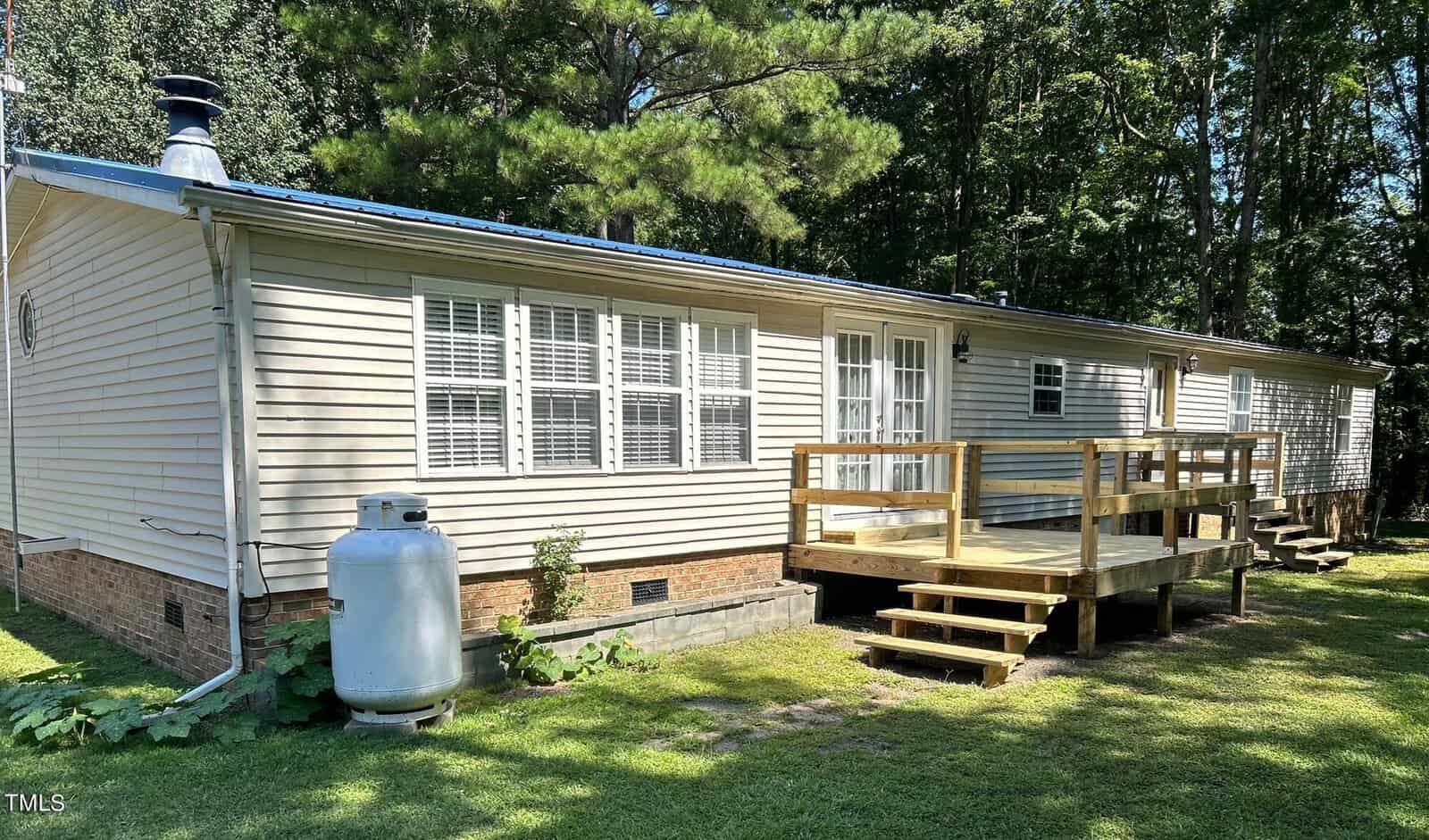 A beige mobile home with a small wooden deck and steps leading to the entrance. The home has several windows and is surrounded by green grass and tall trees. A propane tank is visible on the side.