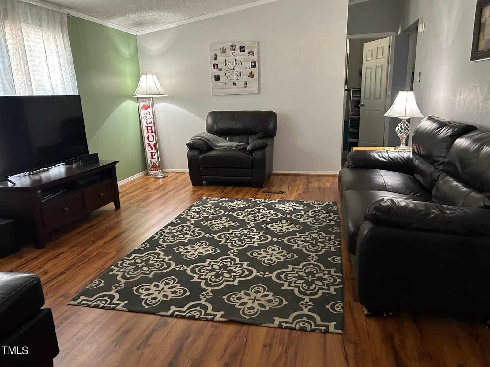 A cozy living room with dark leather sofas, a patterned rug, and wooden floors. There are two tall lamps beside the sofas, a flat-screen TV on a stand, and wall decor with family photos and the word "Home.