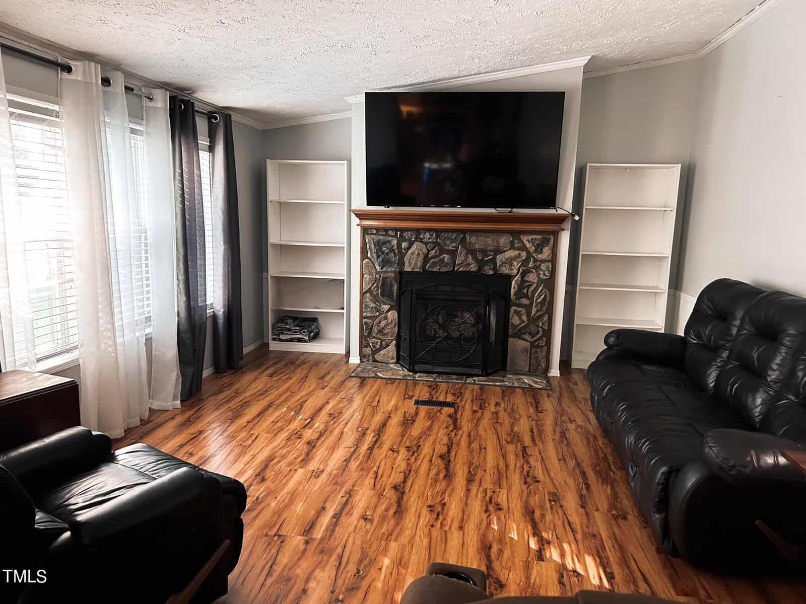 A cozy living room with hardwood floors, a stone fireplace, a large TV mounted above it, and two leather recliners. There are built-in shelves on either side of the fireplace and windows with sheer curtains letting in natural light.