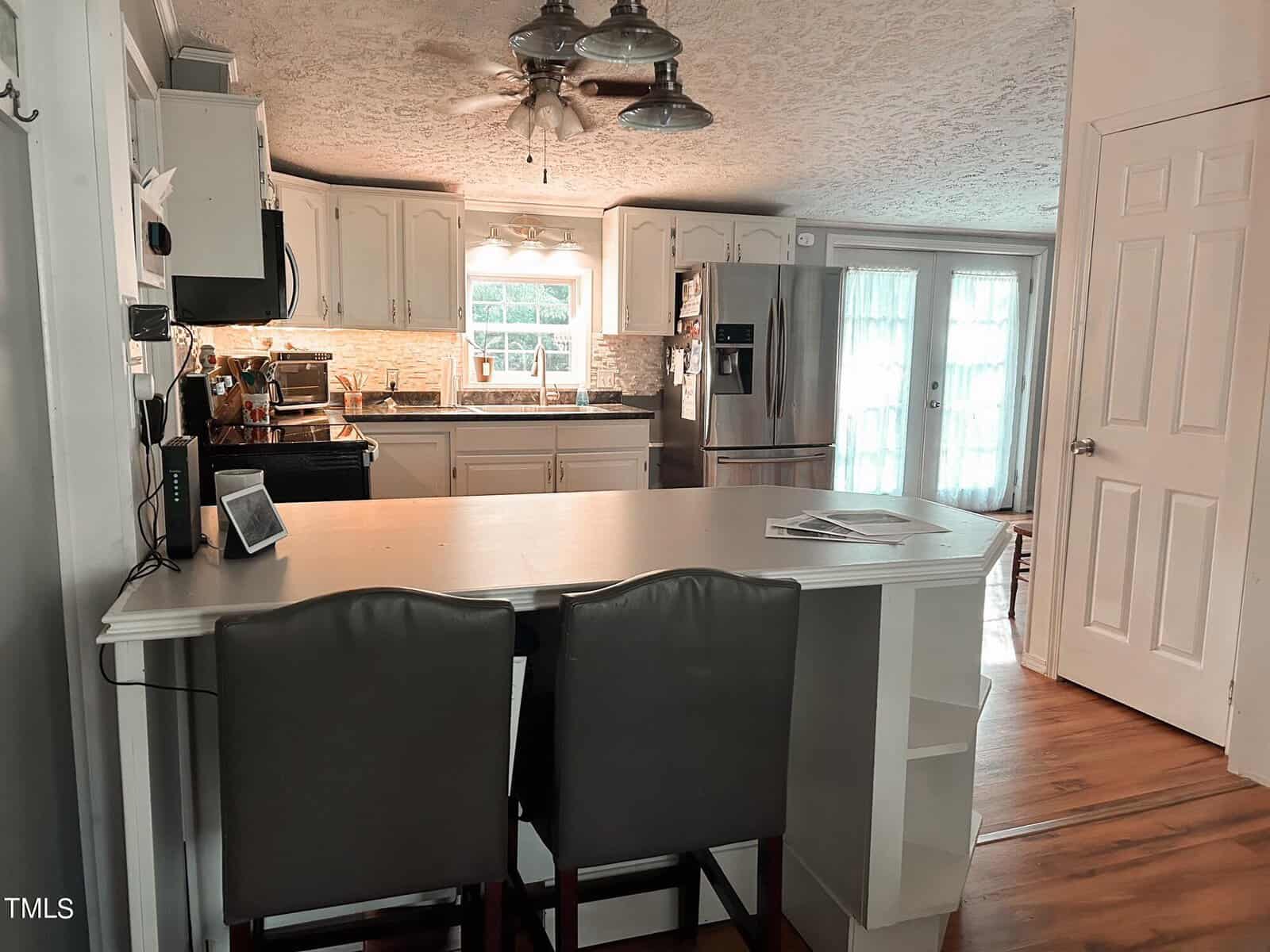 A cozy kitchen with white cabinets, a dark countertop, and stainless steel appliances. There's a central island with two chairs, a ceiling fan above, and double doors leading outside. Light streams in from a window and the doors.