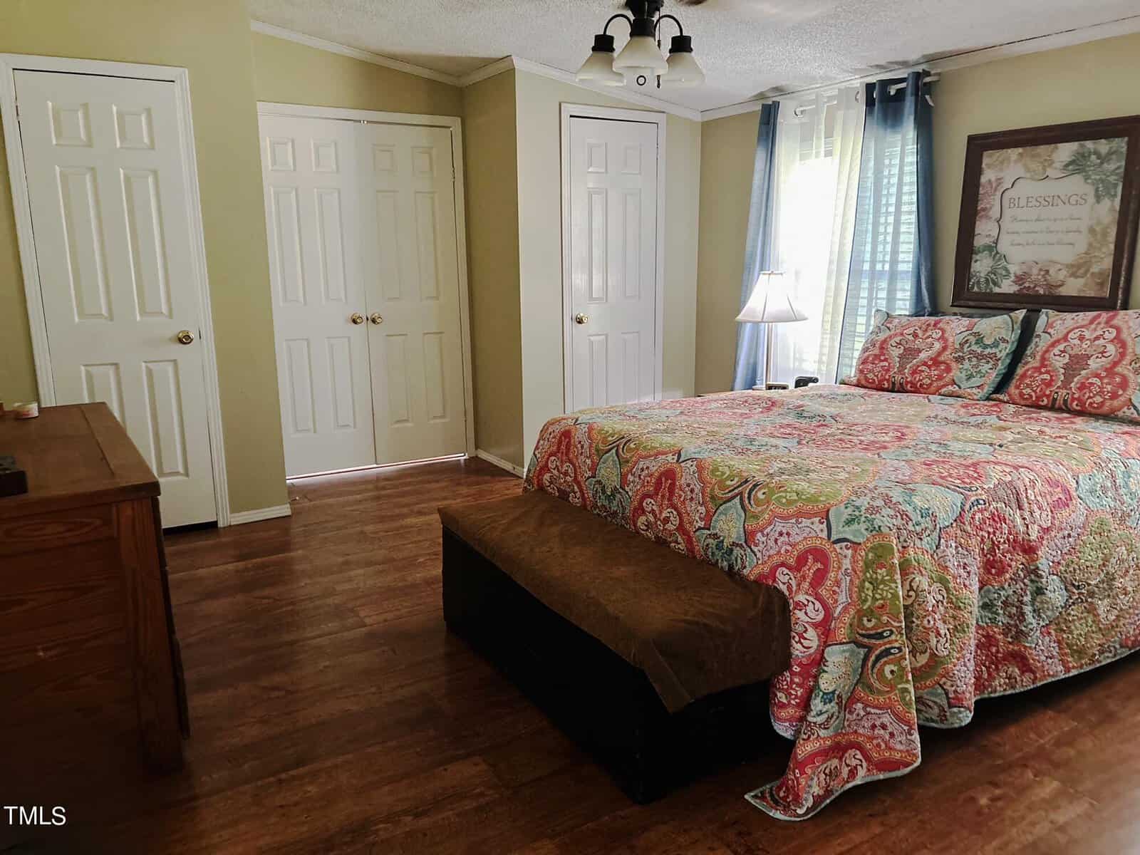 A cozy bedroom with a colorful bedspread, wooden floors, and light green walls. There's a framed artwork on the wall, a window with blue curtains, and a closet with white doors. A small lamp is on the bedside table.