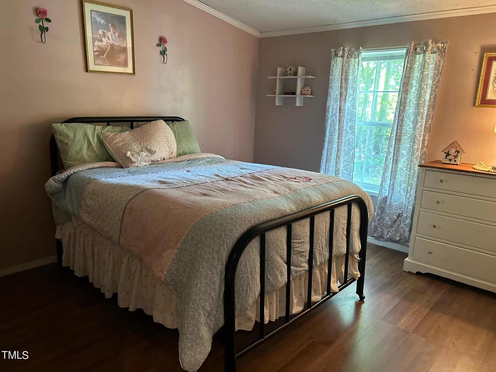 A cozy bedroom with a neatly made bed featuring a pastel quilt and decorative pillows. Walls are painted in soft pink, adorned with a wall shelf and framed art. A window with sheer curtains allows natural light to fill the room, highlighting a white dresser.