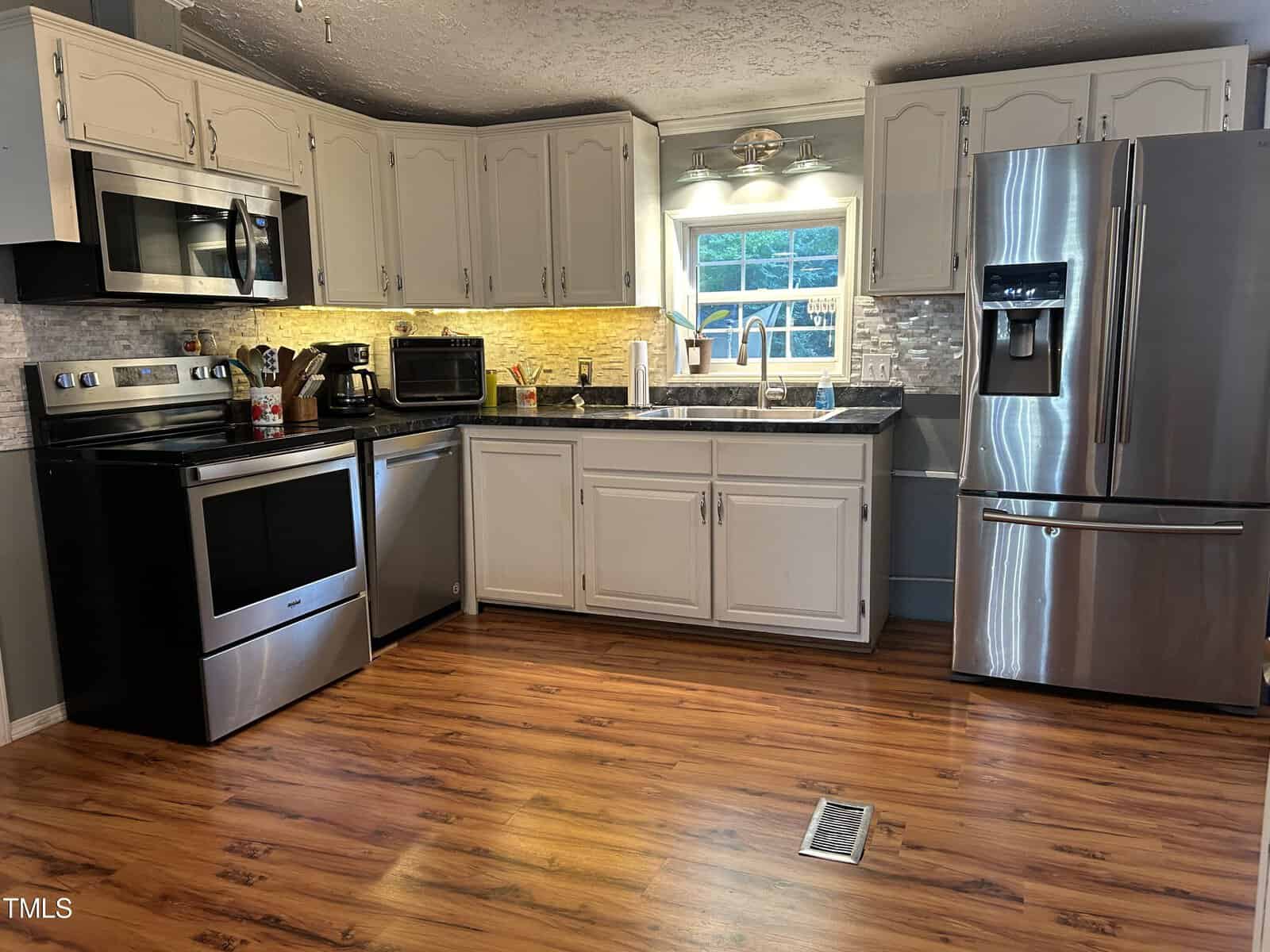 A modern kitchen with stainless steel appliances, including a refrigerator and stove. Light-colored cabinets with a black countertop surround a sink under a window. The floor is hardwood, and a tile backsplash completes the look.