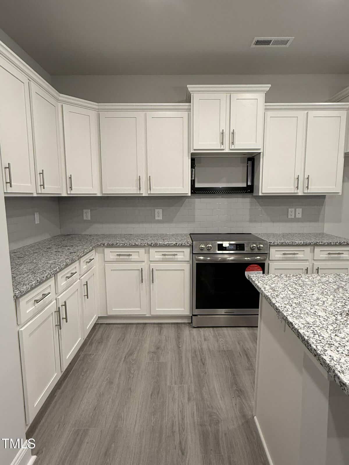 Modern kitchen with white cabinets, granite countertops, light gray subway tile backsplash, stainless steel oven, light wood flooring, and minimal decor. The space is clean, bright, and uncluttered.