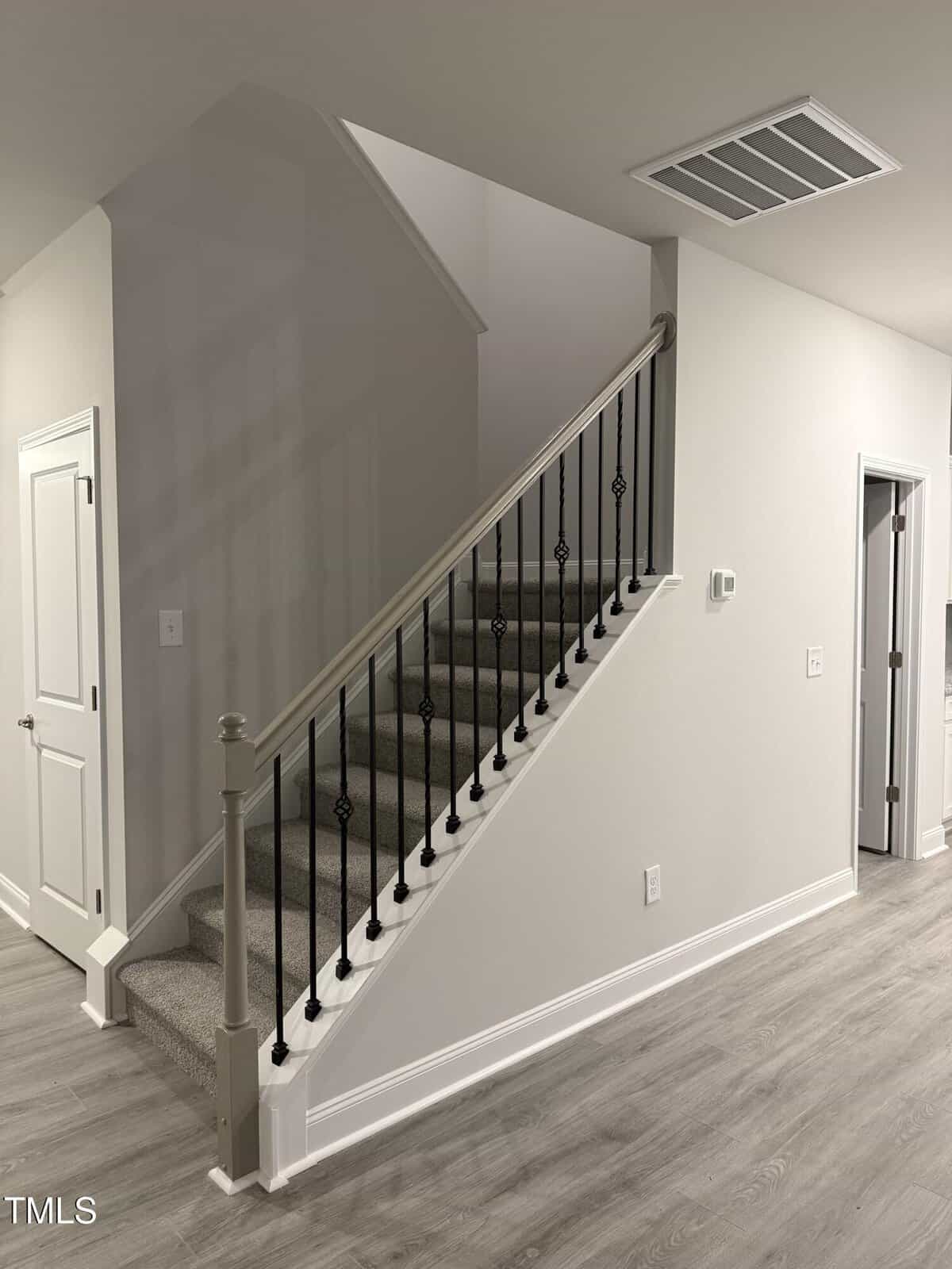 A carpeted staircase with black metal railings and beige handrails in a modern home with light gray walls and wood-style flooring. Several doors lead off the hallway, and an air vent is visible on the ceiling.