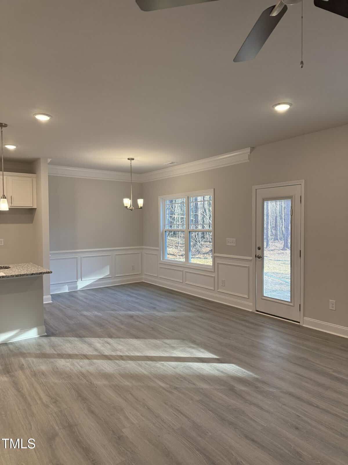 Bright, empty dining and kitchen area with light wood flooring, large windows, a glass door leading outside, white trim and cabinetry, and a ceiling fan with lights. Natural light fills the room.