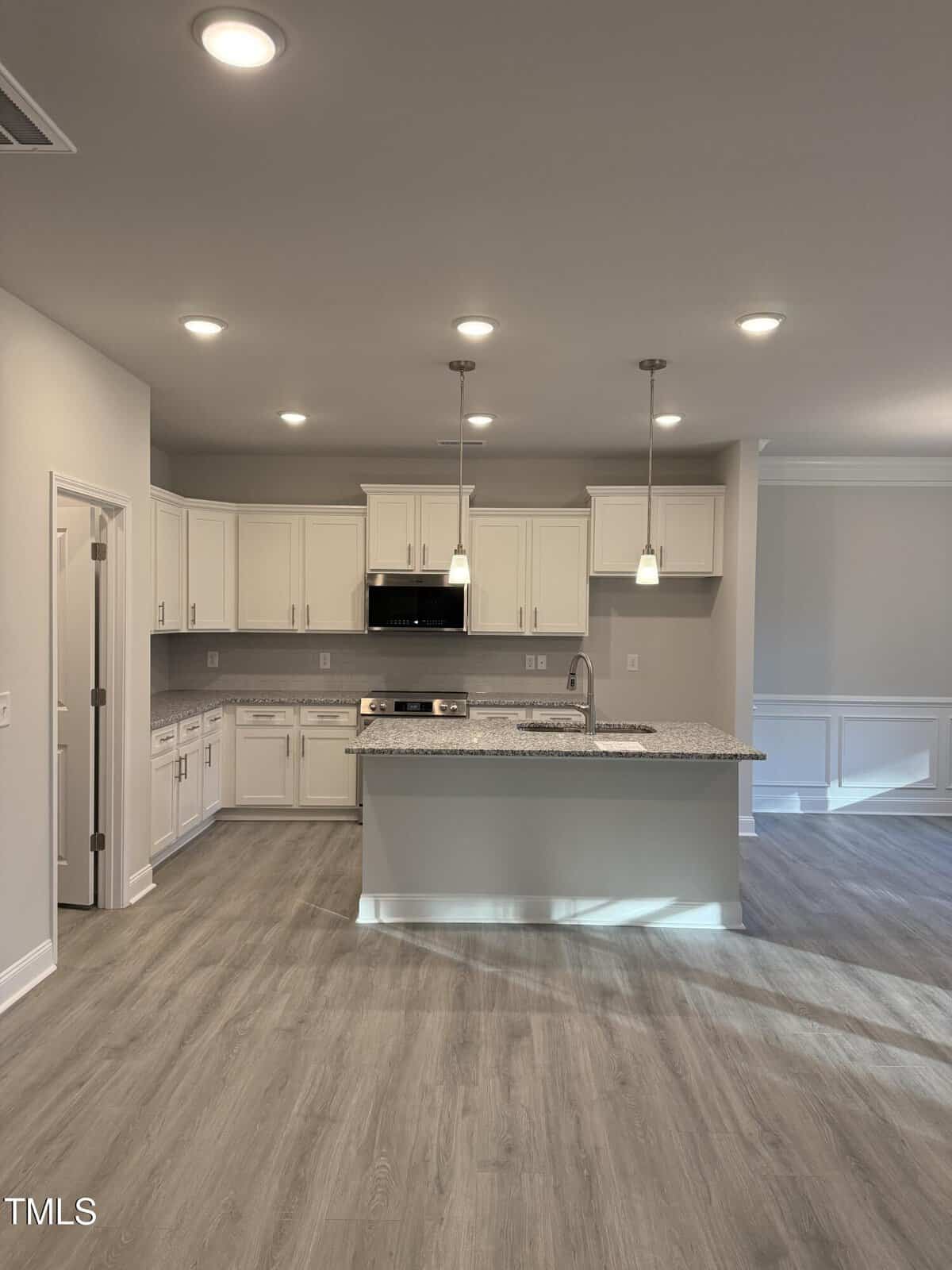 Modern kitchen with white cabinets, stainless steel appliances, granite island with sink, pendant lights, and wood-style flooring. Recessed ceiling lights brighten the open layout with neutral walls and wainscoting in the adjacent room.
