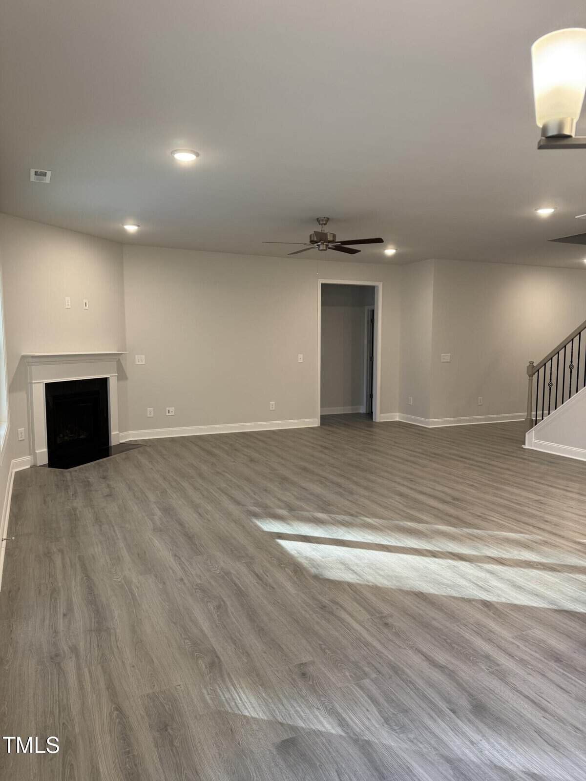 Spacious living room with light gray wood flooring, neutral walls, recessed lighting, a ceiling fan, a corner fireplace, and a view of a staircase with metal railing. Sunlight streams in from a window out of frame.