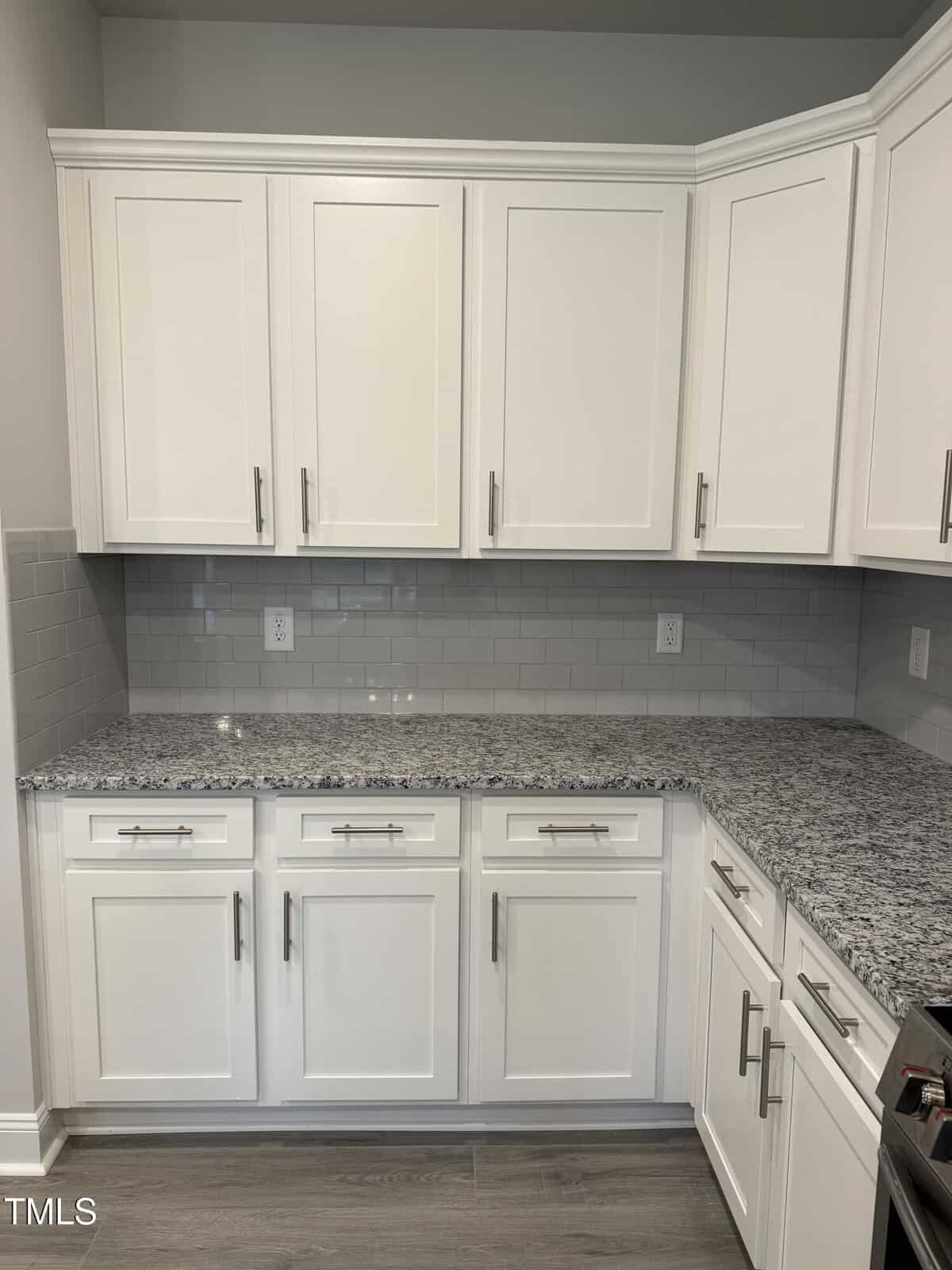 A modern kitchen features white cabinets, silver handles, a gray subway tile backsplash, speckled granite countertops, and gray wood-look flooring. Multiple electrical outlets are visible above the counter.