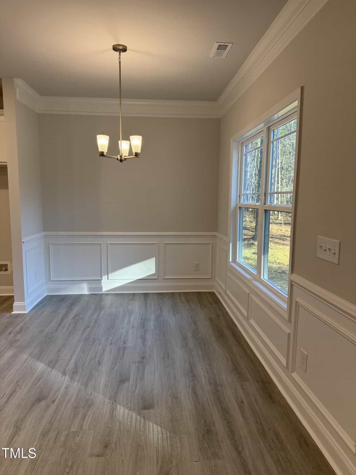 A bright, empty dining room with beige walls, white wainscoting, light wood flooring, a modern chandelier, and large windows overlooking a wooded area.