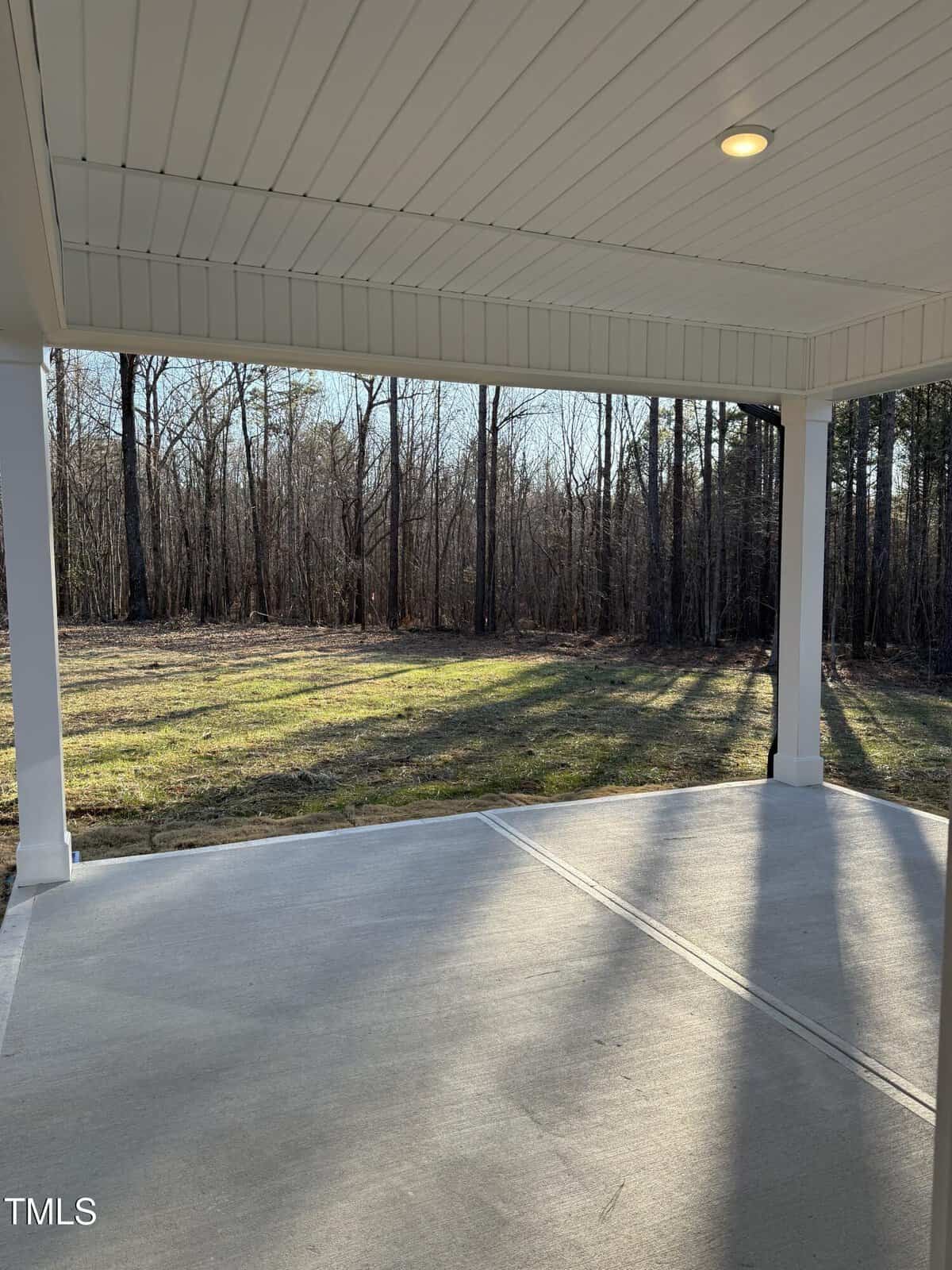 A covered concrete patio with a view of a grassy yard bordered by leafless trees. Sunlight casts long shadows from the trees onto the grass and patio.