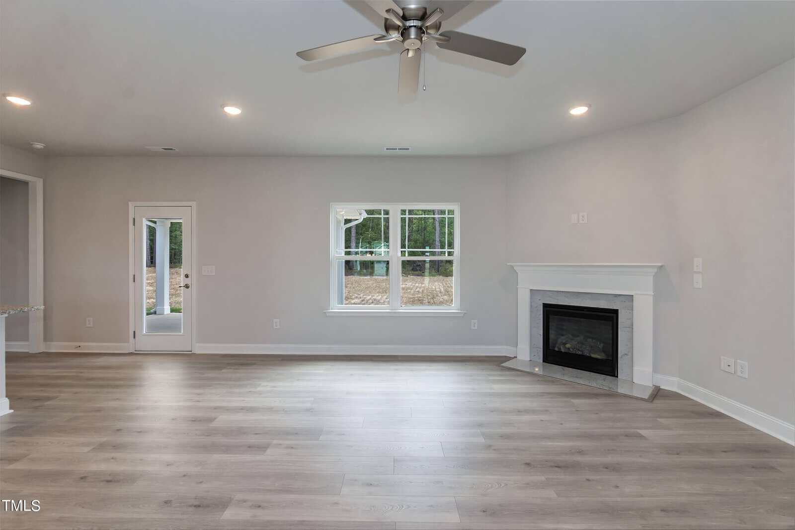 A modern living room with light gray walls, wide plank wood flooring, a ceiling fan with lights, a white framed window, a glass door, and a white fireplace with a black firebox.