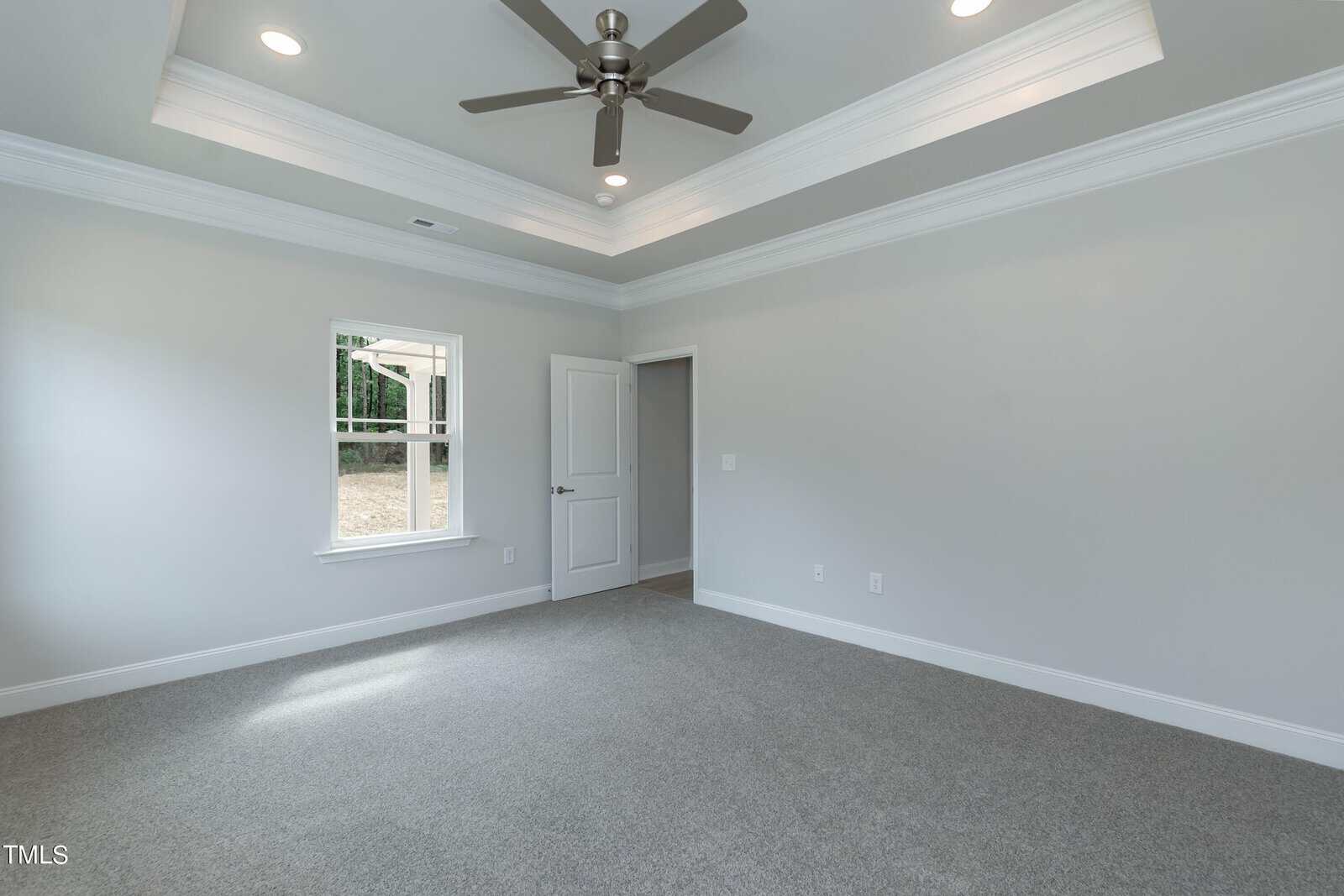 Empty, unfurnished room with gray carpet, light gray walls, white trim, a ceiling fan with lights, tray ceiling, one window, and an open door leading to another area. Natural light enters through the window.