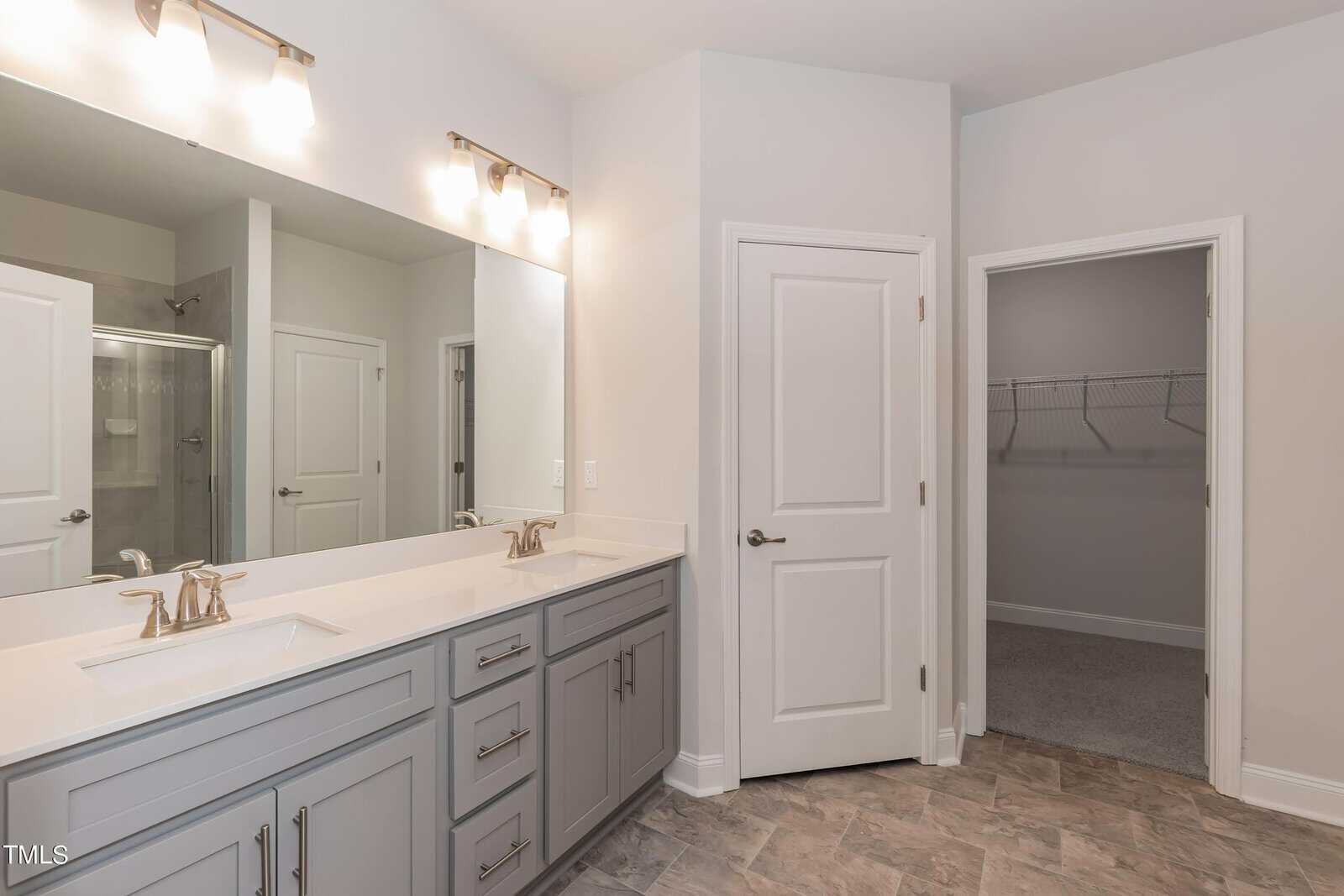 Modern bathroom with double sinks, gray cabinets, large mirror, and bright lighting. A door opens to a walk-in closet with shelves. Neutral tile and wall colors create a clean, spacious look.