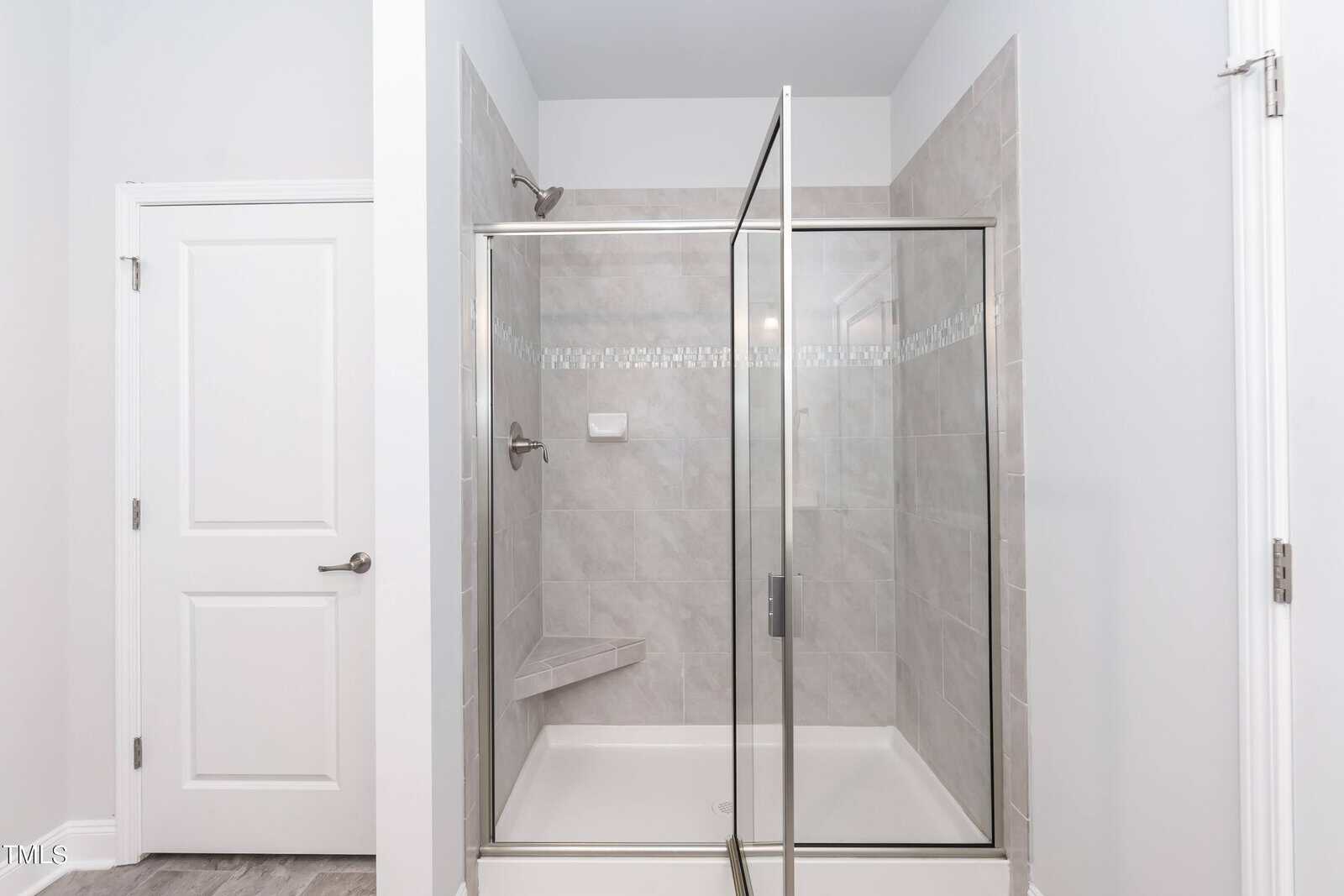A modern bathroom features a glass-enclosed shower with beige tile, a built-in corner bench, and a white door to the left. The walls and ceiling are painted white.