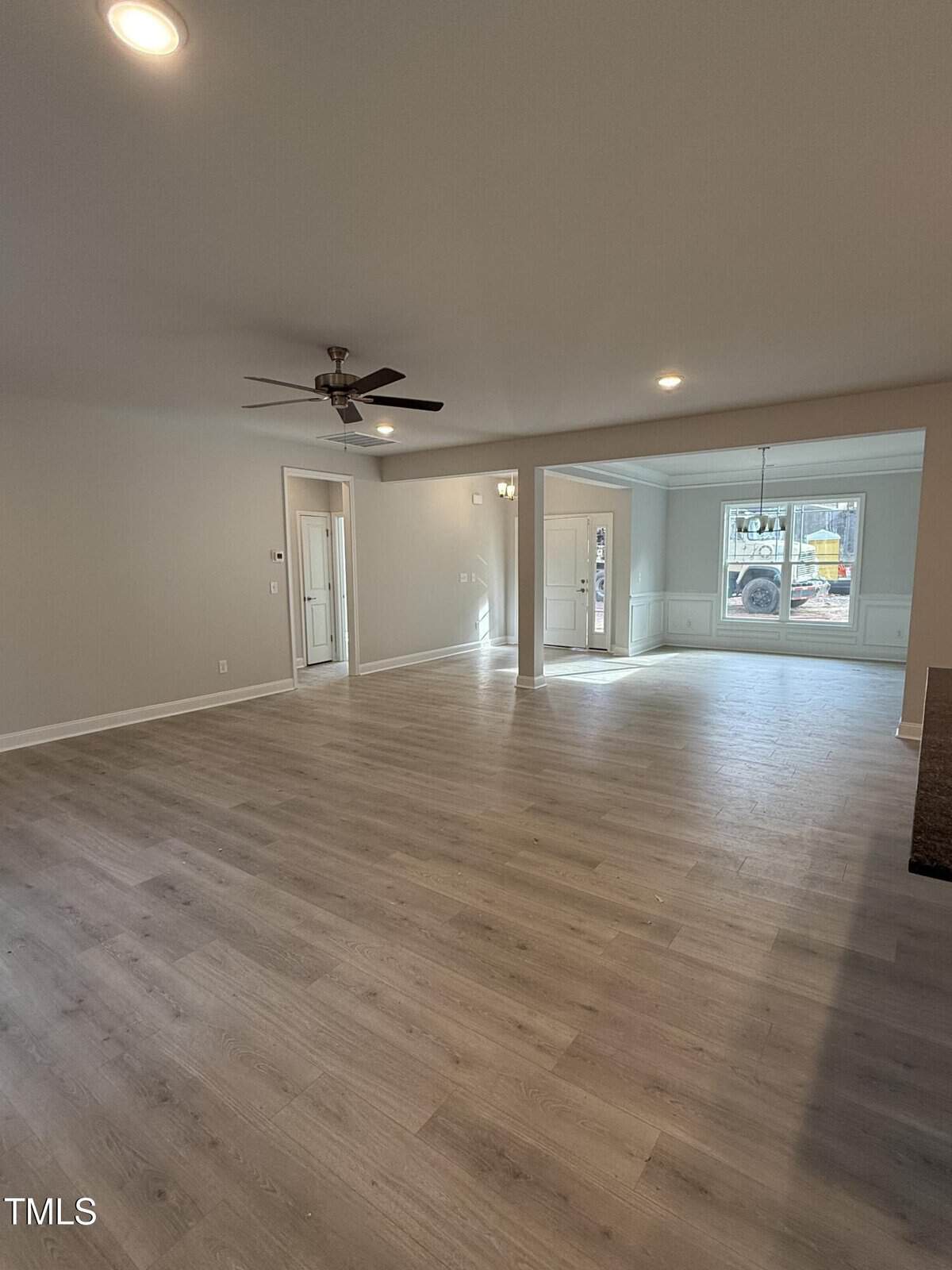 Spacious empty living area with light wood floors, light gray walls, a ceiling fan, and recessed lighting. Large front window lets in natural light. Open layout connects to another room in the background.
