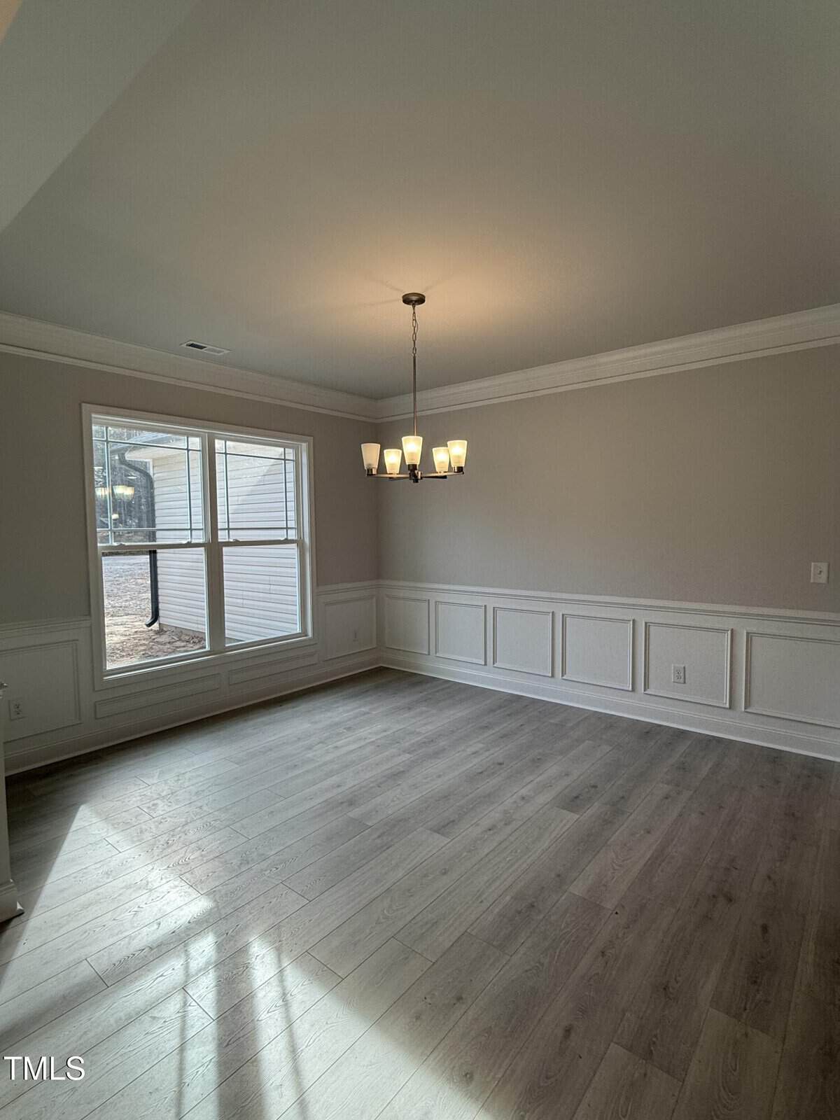 A bright, empty room with light wood flooring, beige walls with white wainscoting, a large window letting in natural light, and a modern chandelier hanging from the ceiling.
