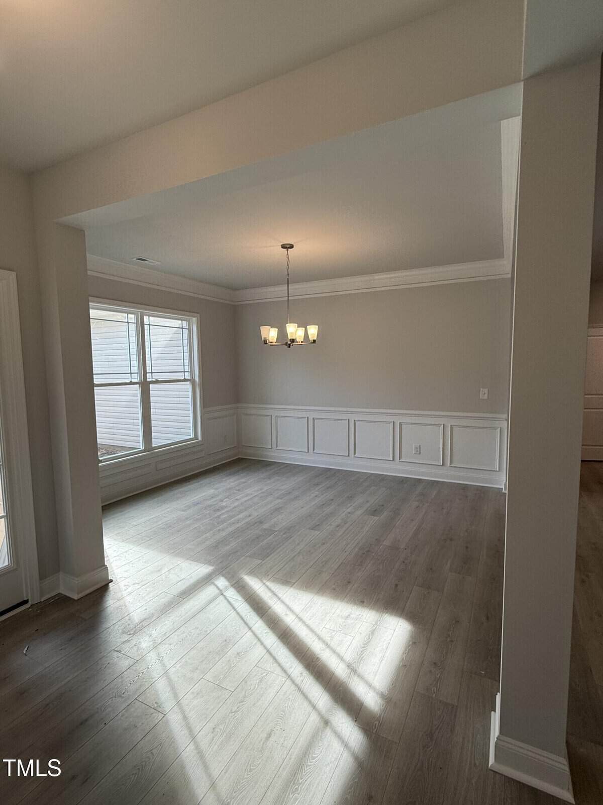 A bright, empty dining room with light wood flooring, beige walls, white wainscoting, a large window, and a modern chandelier hanging from the ceiling. Sunlight streams in, creating shadows on the floor.