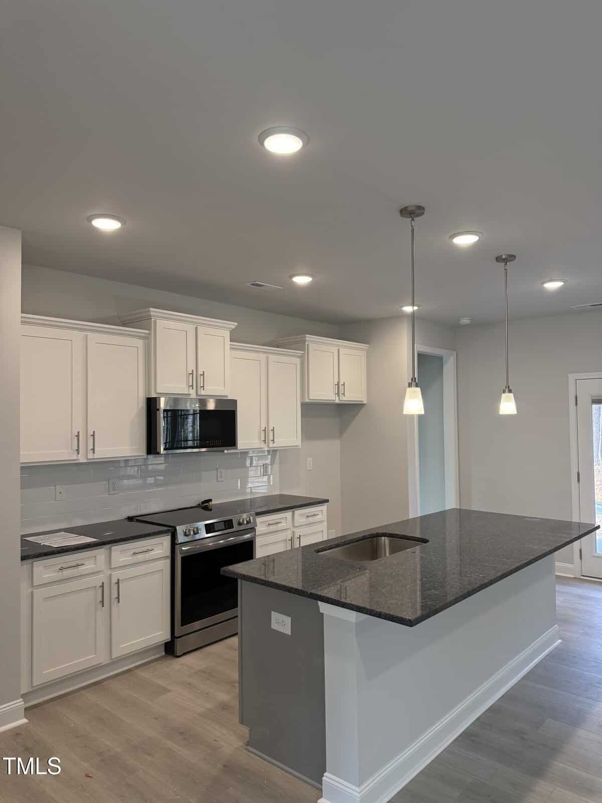 Modern kitchen with white cabinets, stainless steel appliances, black granite island with sink, pendant lights, light wood flooring, and a door leading outside. Recessed ceiling lights brighten the space.