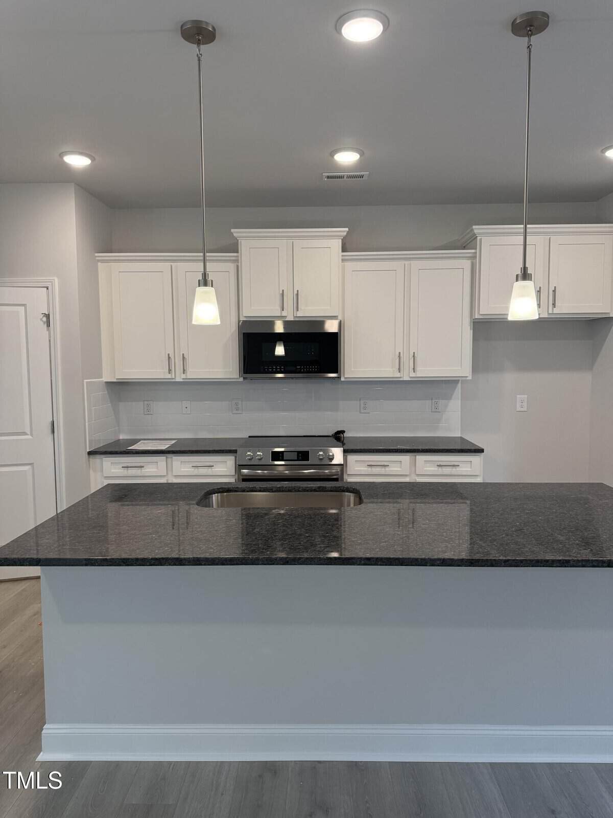 Modern kitchen with white cabinets, stainless steel appliances, black granite countertops, a center island with sink, two pendant lights above, and light gray walls and flooring.