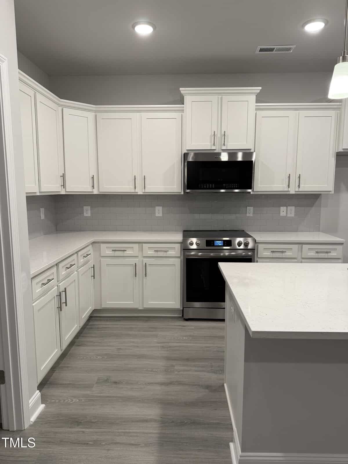 Modern kitchen with white cabinets, stainless steel oven and microwave, light gray tile backsplash, white quartz countertops, and light gray wood flooring. An island is visible in the foreground under pendant lighting.