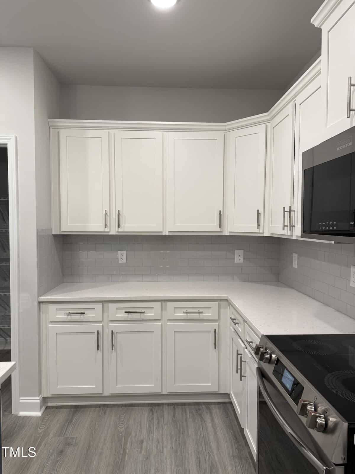 Modern kitchen with white cabinets, light gray subway tile backsplash, white countertops, stainless steel oven and microwave, and light wood flooring. Cabinets and drawers have silver handles. Walls are painted light gray.