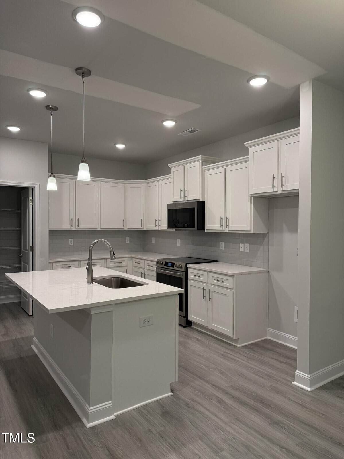 Modern kitchen with white cabinets, stainless steel appliances, an island with a sink and pendant lights, light gray backsplash, and wood-look flooring. There is also a walk-in pantry to the left.