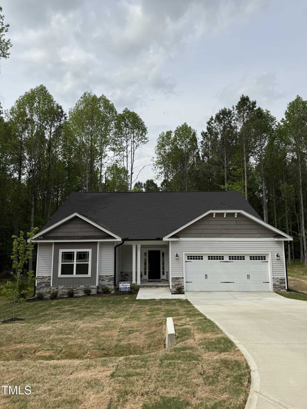 A modern single-story house with gray siding, a dark roof, and a white double garage sits in front of a wooded area. The lawn is newly planted, and a concrete driveway leads to the garage.