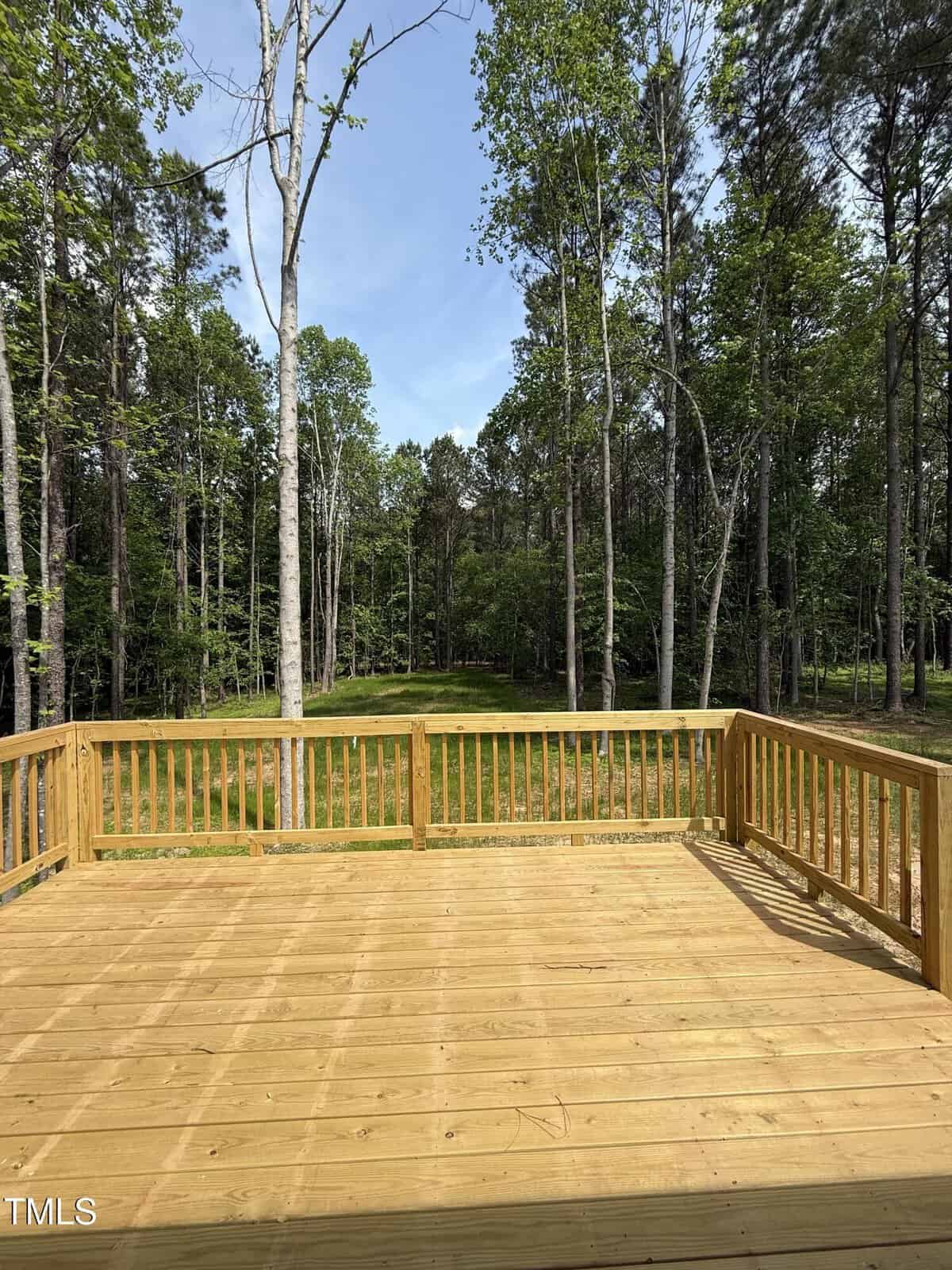 A spacious wooden deck with railings overlooks a grassy yard surrounded by tall trees and dense forest under a clear blue sky.