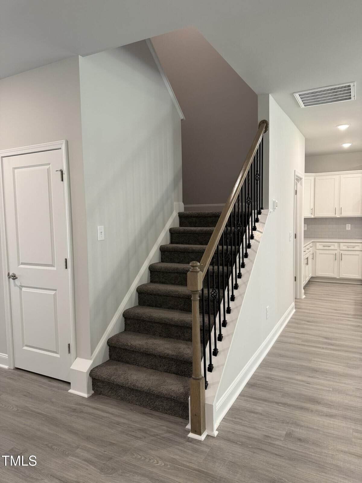 A modern interior featuring carpeted stairs with a wooden handrail and black spindles. To the left is a closed white door, and to the right, a hallway leads to a bright kitchen with white cabinets. Light gray walls and flooring.