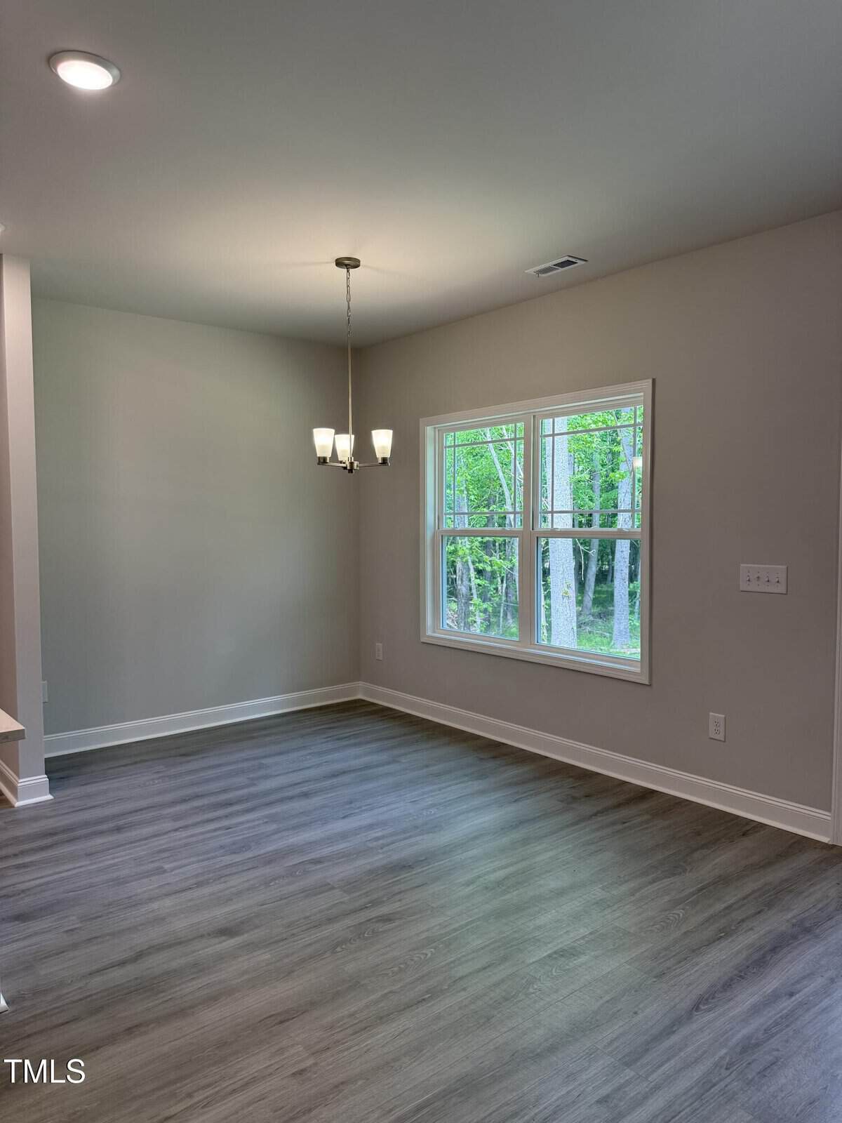 A bright, empty dining area with light wood flooring, beige walls, a modern chandelier, and a large window showing green trees outside.