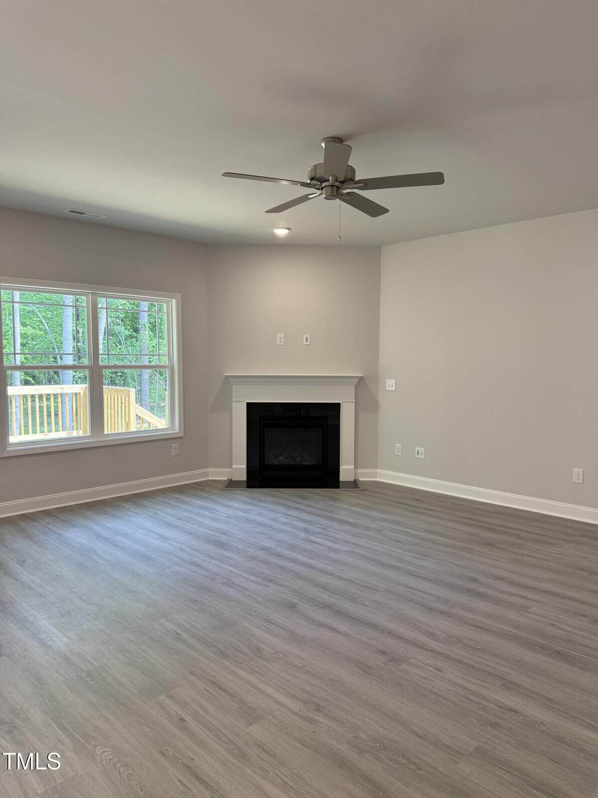 A bright, empty living room with light gray walls, wood-style flooring, a ceiling fan, a fireplace, and large windows overlooking a deck and green trees outside.