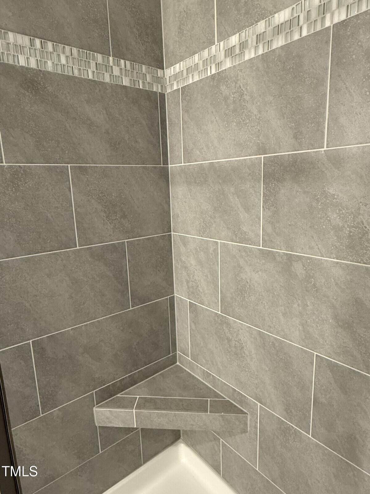 A tiled shower corner with gray rectangular tiles, a small triangular corner shelf, and a decorative strip of mosaic tiles near the top.