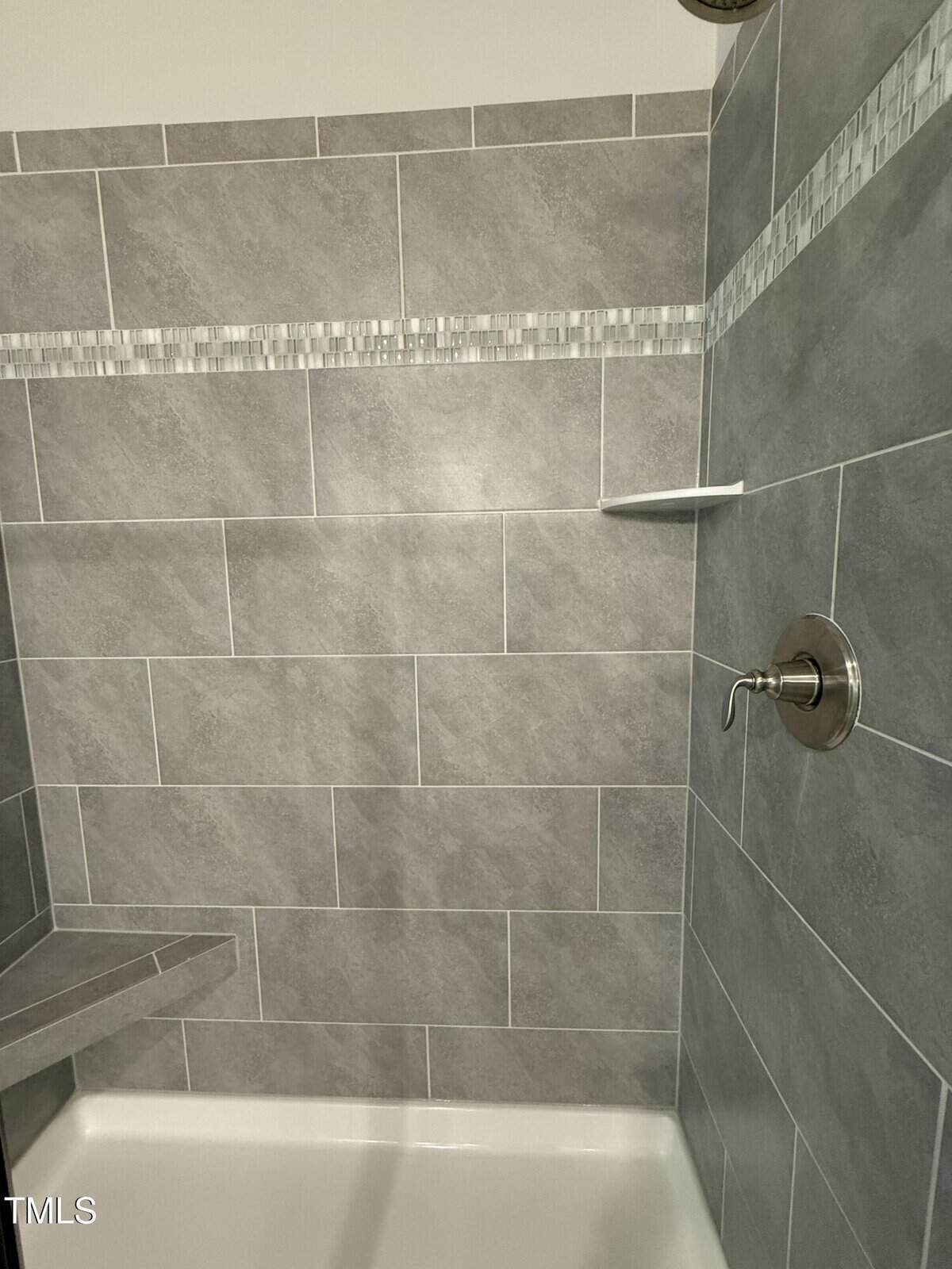 Shower with gray rectangular tiles, a decorative horizontal strip, a metal showerhead, a round control knob, a corner seat, and a small wall shelf.