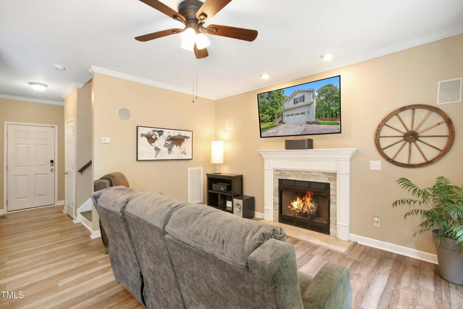 A cozy living room with a beige color scheme features a gray couch, a ceiling fan, and a fireplace beneath a wall-mounted TV. A map and wagon wheel decor adorn the walls. Natural light streams through a window, illuminating a potted plant.
