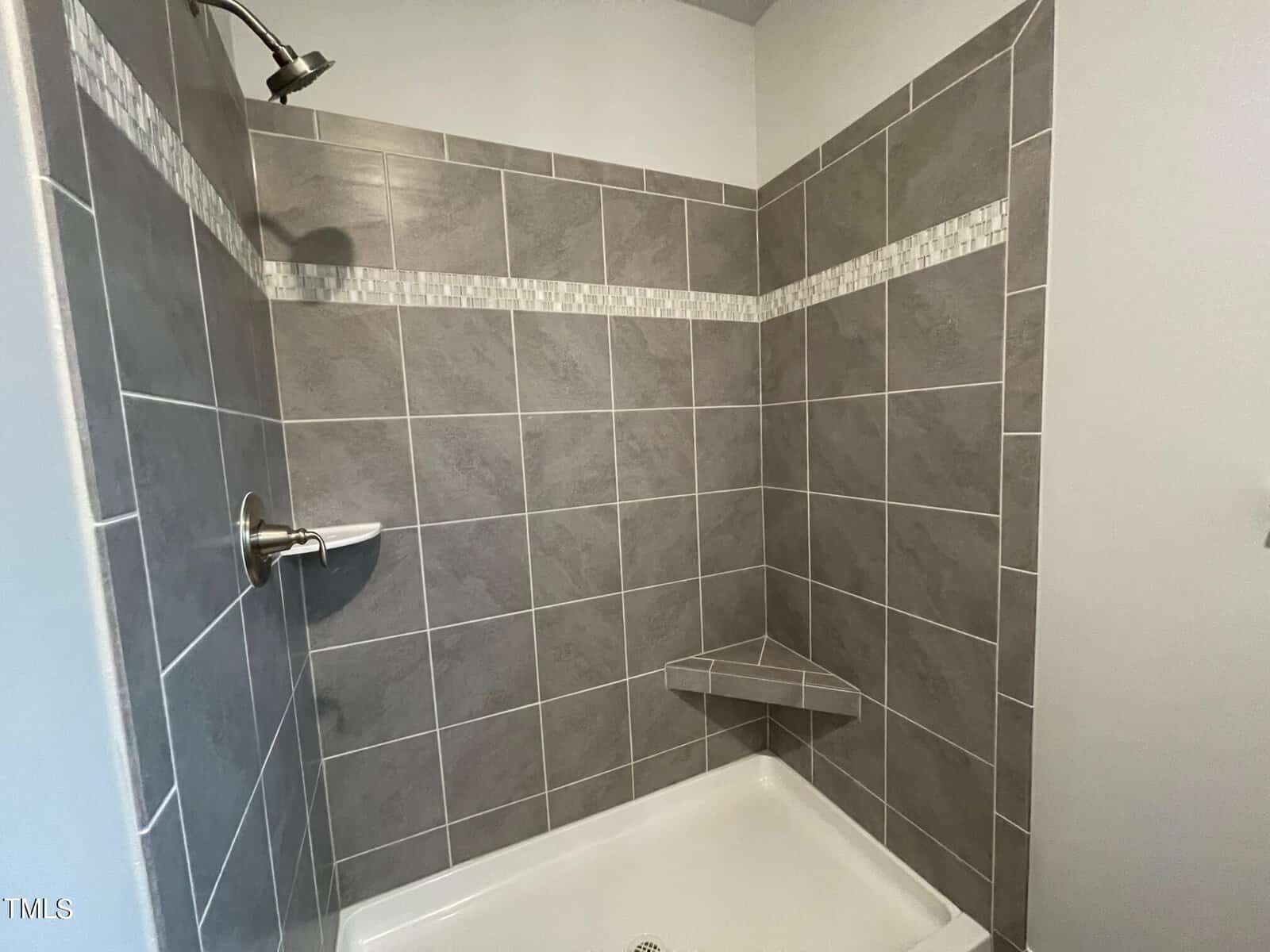 A modern shower with gray rectangular tiles, a decorative horizontal accent strip, a silver showerhead and faucet, and a small triangular corner shelf.