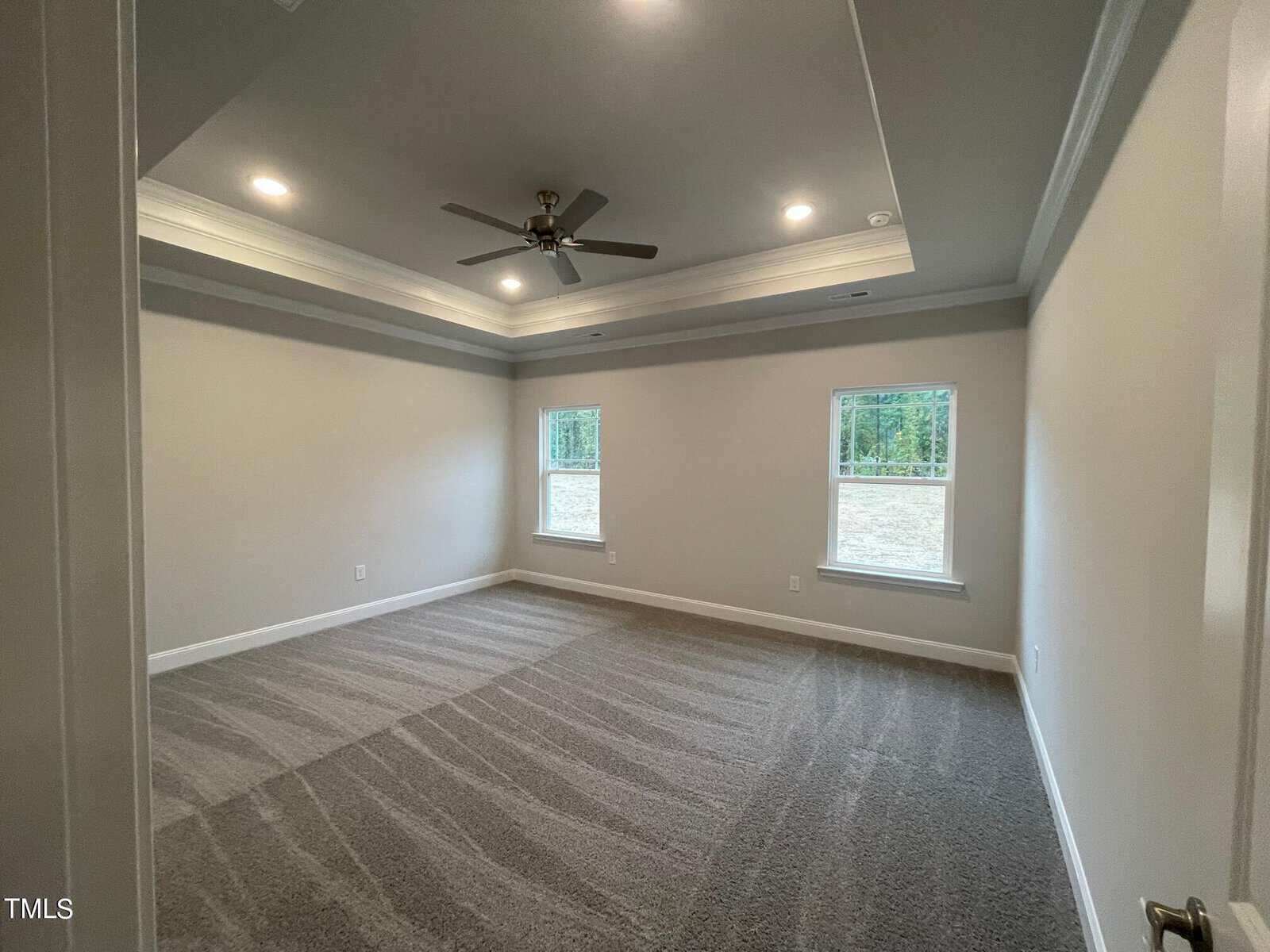A spacious, empty room with light gray walls, two large windows, carpeted floor, recessed lighting, and a ceiling fan with tray ceiling detailing. Natural light enters through the windows.