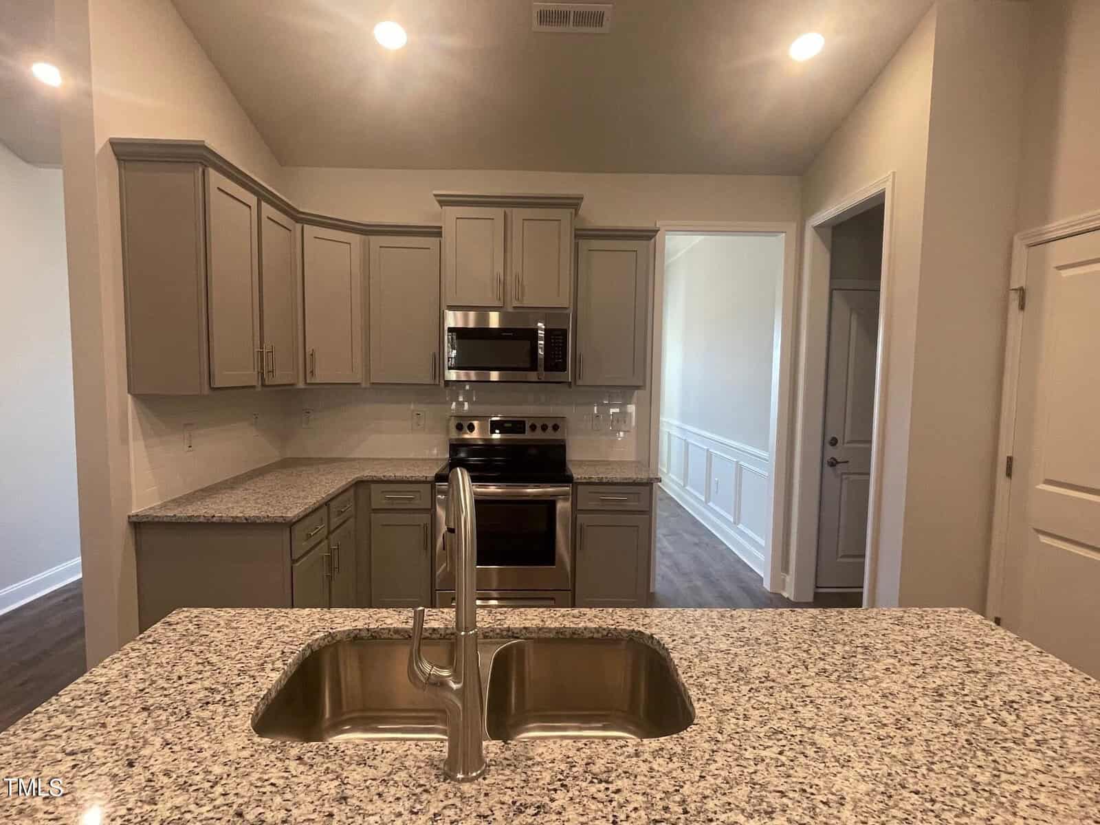Modern kitchen with granite countertops, a double sink, stainless steel appliances including a microwave and oven, gray cabinets, and tile backsplash. The room is well-lit with recessed ceiling lights.