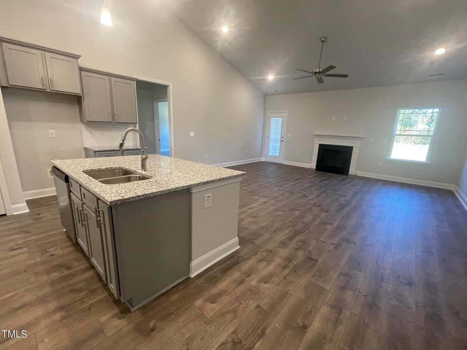 A spacious open-concept living area features wood-style flooring, a modern kitchen with gray cabinets and an island with a double sink, a ceiling fan, a fireplace, and large windows for natural light.