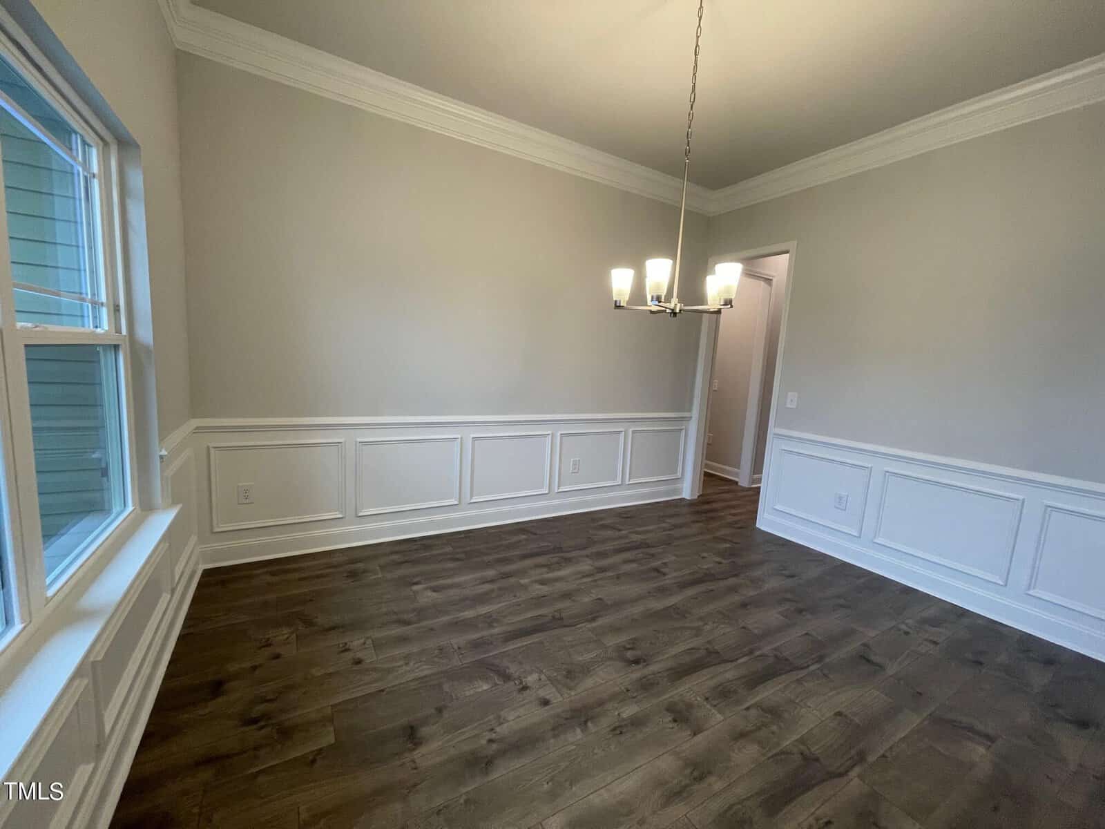 A bright, empty dining room with wood floors, white wainscoting, light gray walls, a window on the left, a modern chandelier, and a doorway leading to another space.