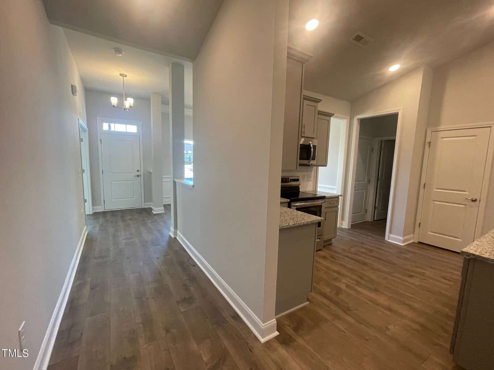Wide hallway in a modern home with wood flooring, light beige walls, an entryway with a door and sidelights, visible kitchen area with granite counters, and multiple doorways leading to other rooms.