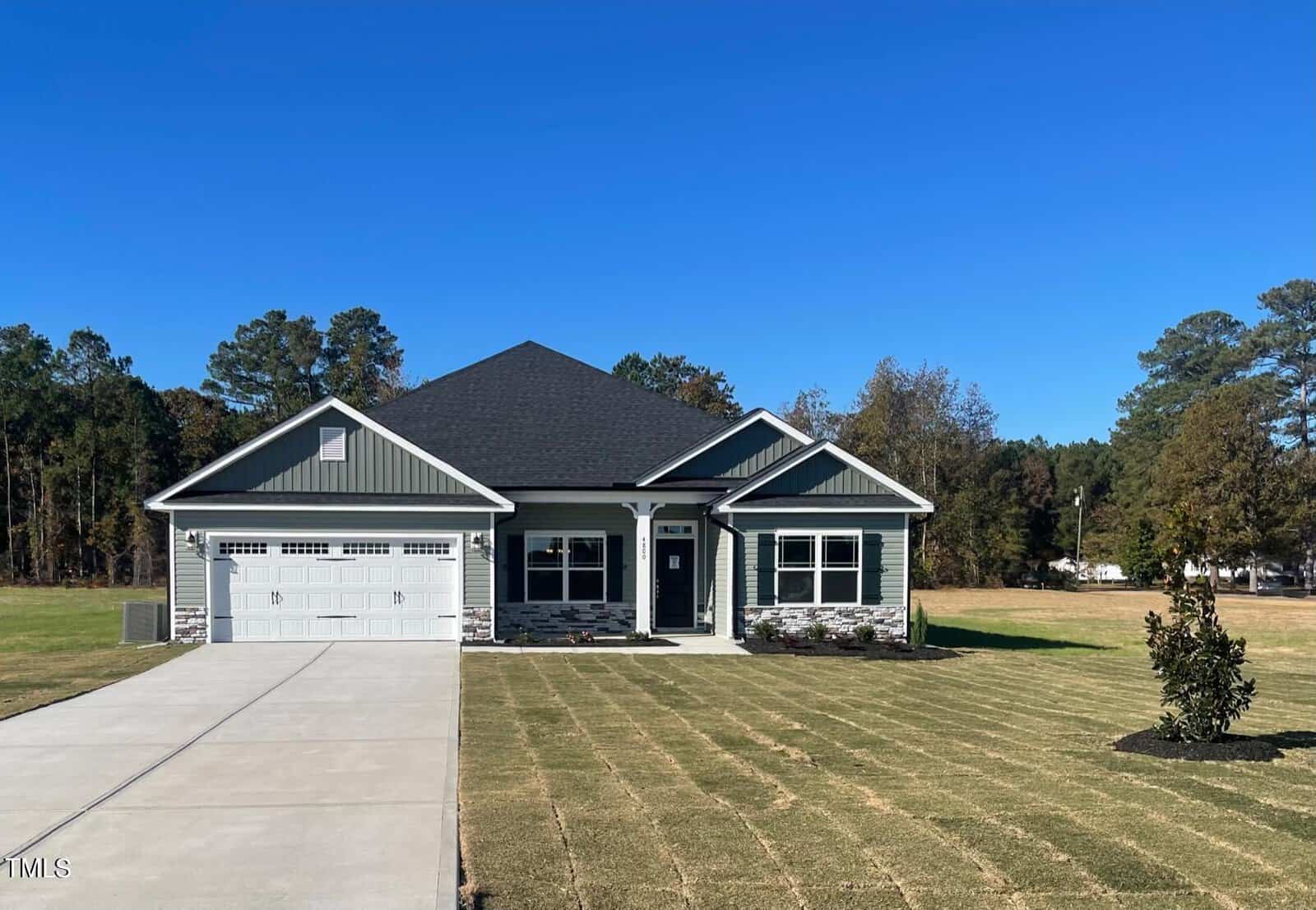 A single-story gray house with white trim, a covered front porch, and a two-car garage sits on a large, freshly landscaped lawn with a concrete driveway. Trees line the background under a clear blue sky.