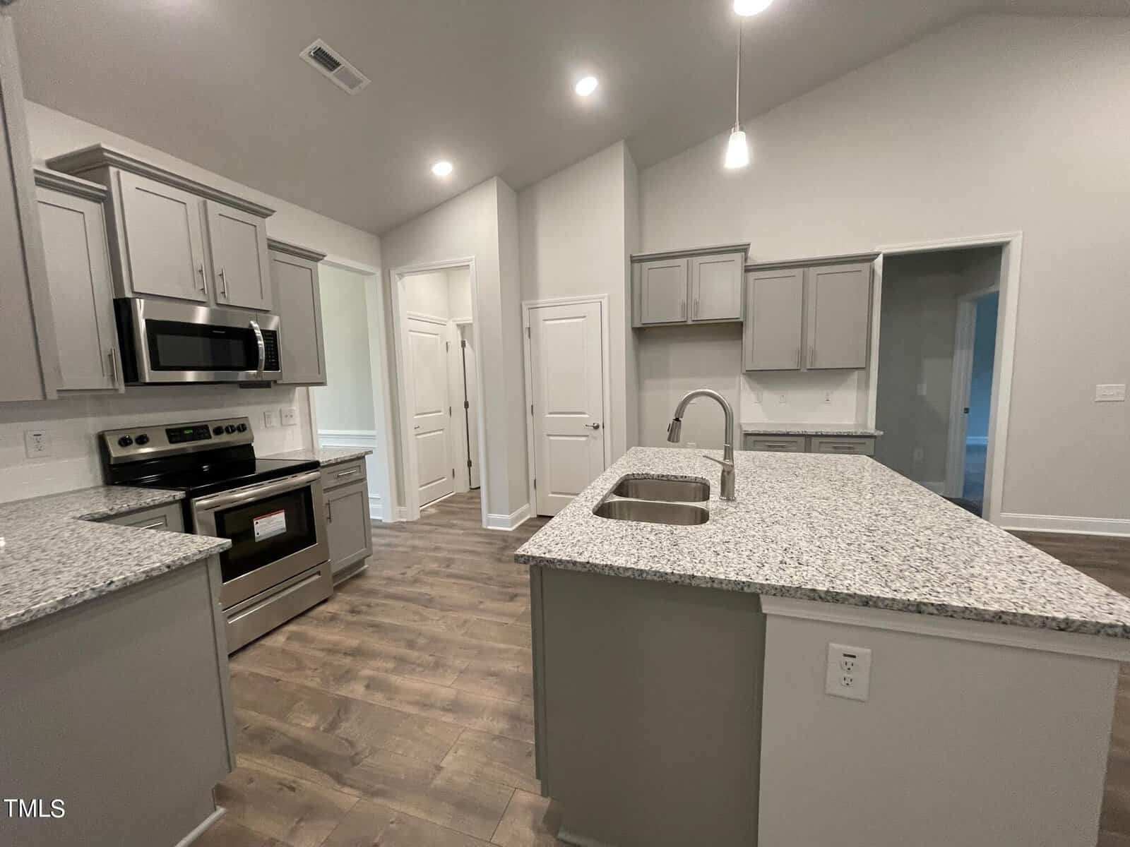 Modern kitchen with gray cabinets, granite countertops, stainless steel appliances, center island with sink, and wood flooring. Bright lighting and vaulted ceiling create an open, spacious feel. Multiple doors lead to other rooms.