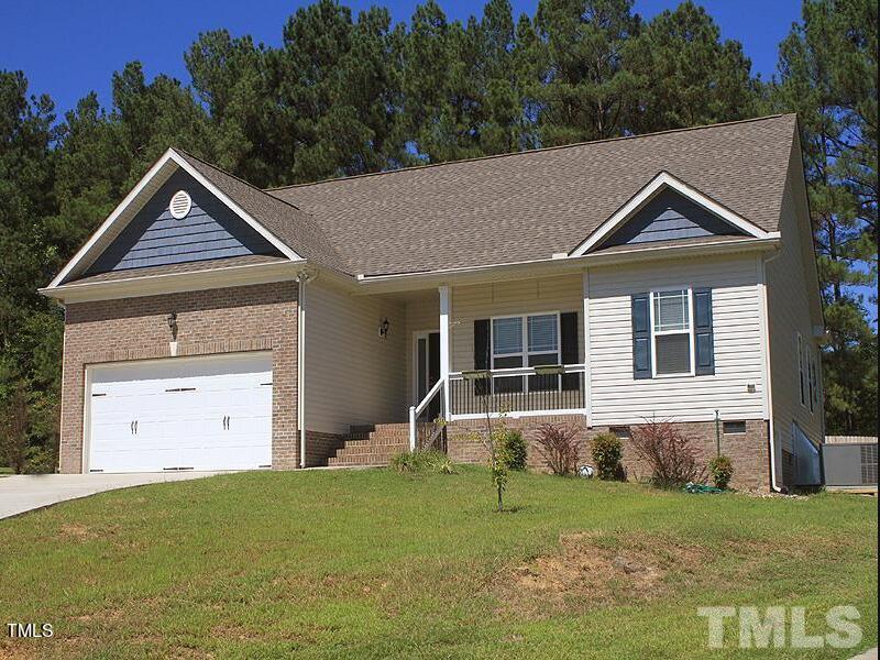 Located at 301 Coachmans Trail, this charming single-story house boasts a brown brick base and beige siding. It features a two-car garage, a cozy front porch, and a sloped driveway, all nestled against a backdrop of tall pine trees under a clear blue sky.
